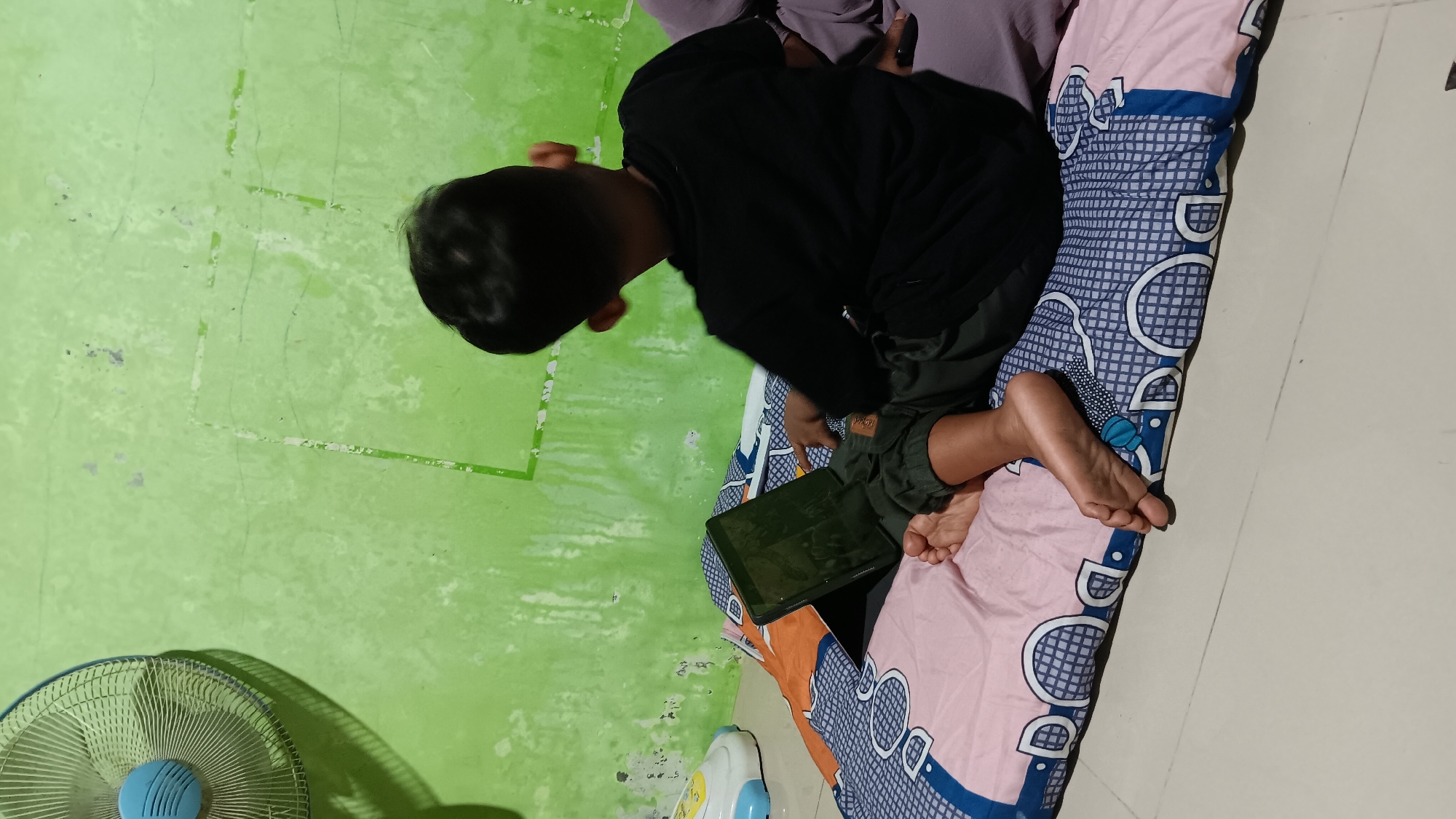 In Sonobekel, Indonesia, a young boy is seen from behind, sitting on a pink and blue patterned mattress, engrossed in a tablet. He is wearing a black t-shirt and dark pants. His bare feet are visible, resting on the mattress. To the left of the boy, a portion of a blue and white electric fan is visible. The background is a green, textured wall with faint white markings that resemble squares or rectangles. The floor appears to be tiled.