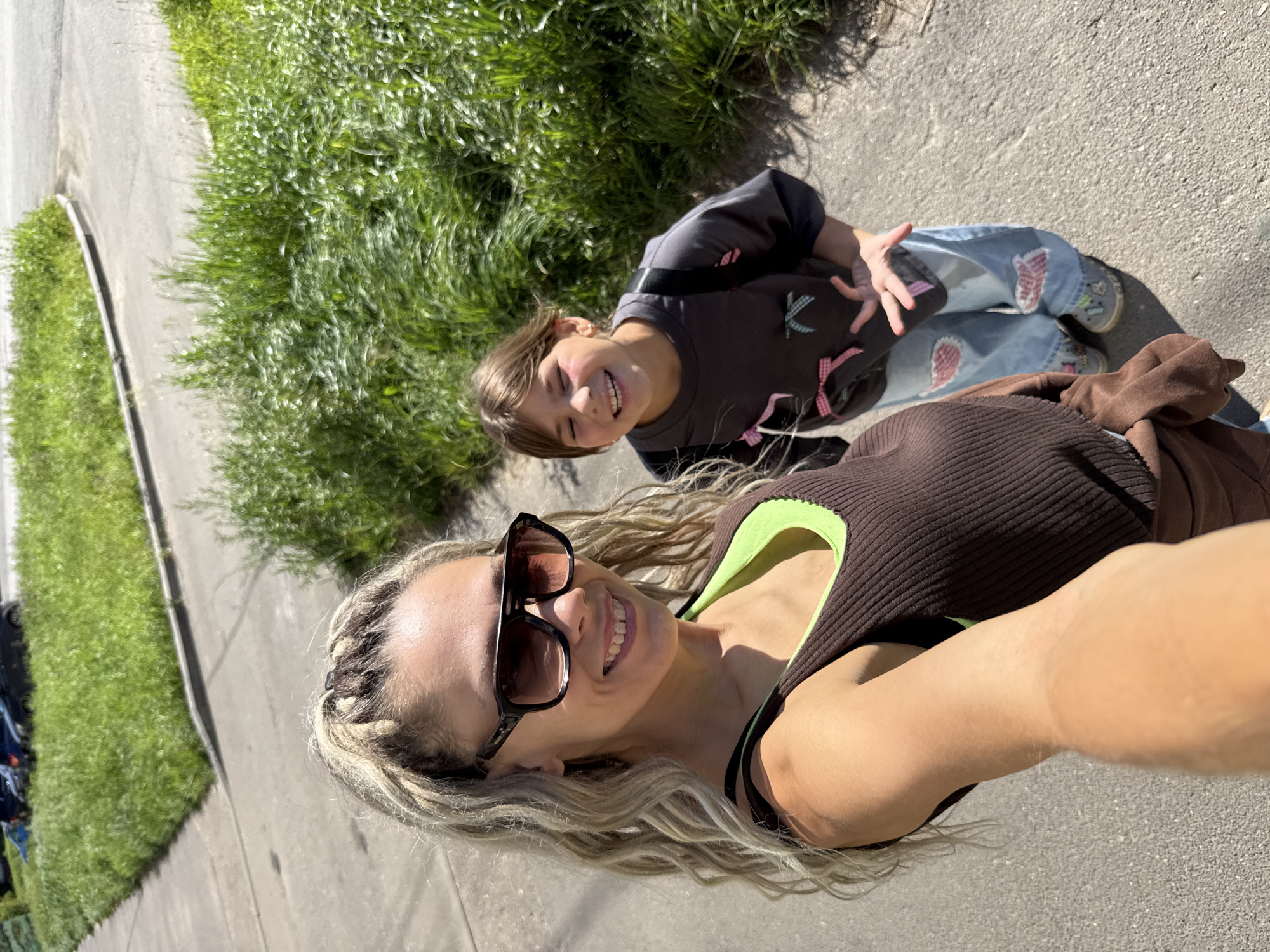 This is a selfie taken outdoors on a sunny day, likely in the city of Rusanovo, Russia. Two individuals, a woman and a girl, are visible. The woman is in the foreground, holding the camera. She has blonde, braided hair and is wearing large sunglasses, a brown ribbed tank top with a neon green lining, and a brown cardigan. The girl is positioned behind her and to the right, smiling widely with her eyes partially closed. She is wearing a dark grey t-shirt with colorful ribbon appliques and light blue jeans with patches. Both are standing on a paved surface, possibly a sidewalk or road, with a grassy embankment to their right. In the background, cars are parked on a paved area adjacent to the grass. The overall scene depicts a casual, outdoor moment between the two individuals.