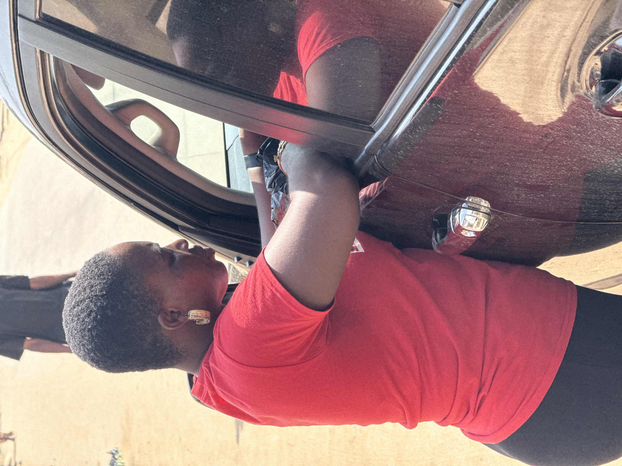 In Abuja, Nigeria, a woman with short, dark hair and large hoop earrings, wearing a red t-shirt and dark leggings, leans into a dark red SUV. She is reaching inside the vehicle, holding a black plastic bag. The woman's face is partially visible as she looks down. The car's window is down, and a hand and arm are visible inside the car, also holding the bag. Another person, wearing a black t-shirt, is partially visible in the background, standing on a sandy surface. The scene appears to be outdoors on a bright, sunny day, with the ground being sandy and dusty. The car is parked, and the overall impression is a transaction or exchange occurring at the vehicle.