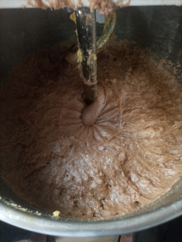 Within the city of Opete, Nigeria, a close-up overhead perspective reveals the interior of a large, polished metal mixing bowl containing a substantial volume of viscous, dark brown batter or dough. A heavy-duty metallic mixing attachment, possibly a dough hook, is partially submerged in the center of the mixture, showing traces of the brown material and some yellowish residue near its upper portion. The batter exhibits distinct textural patterns and swirls, particularly around the mixer attachment, indicative of recent or ongoing mechanical agitation. The upper edge of a white machine housing, likely part of an industrial-scale mixer, is visible along the top perimeter of the frame. No individuals are visible.