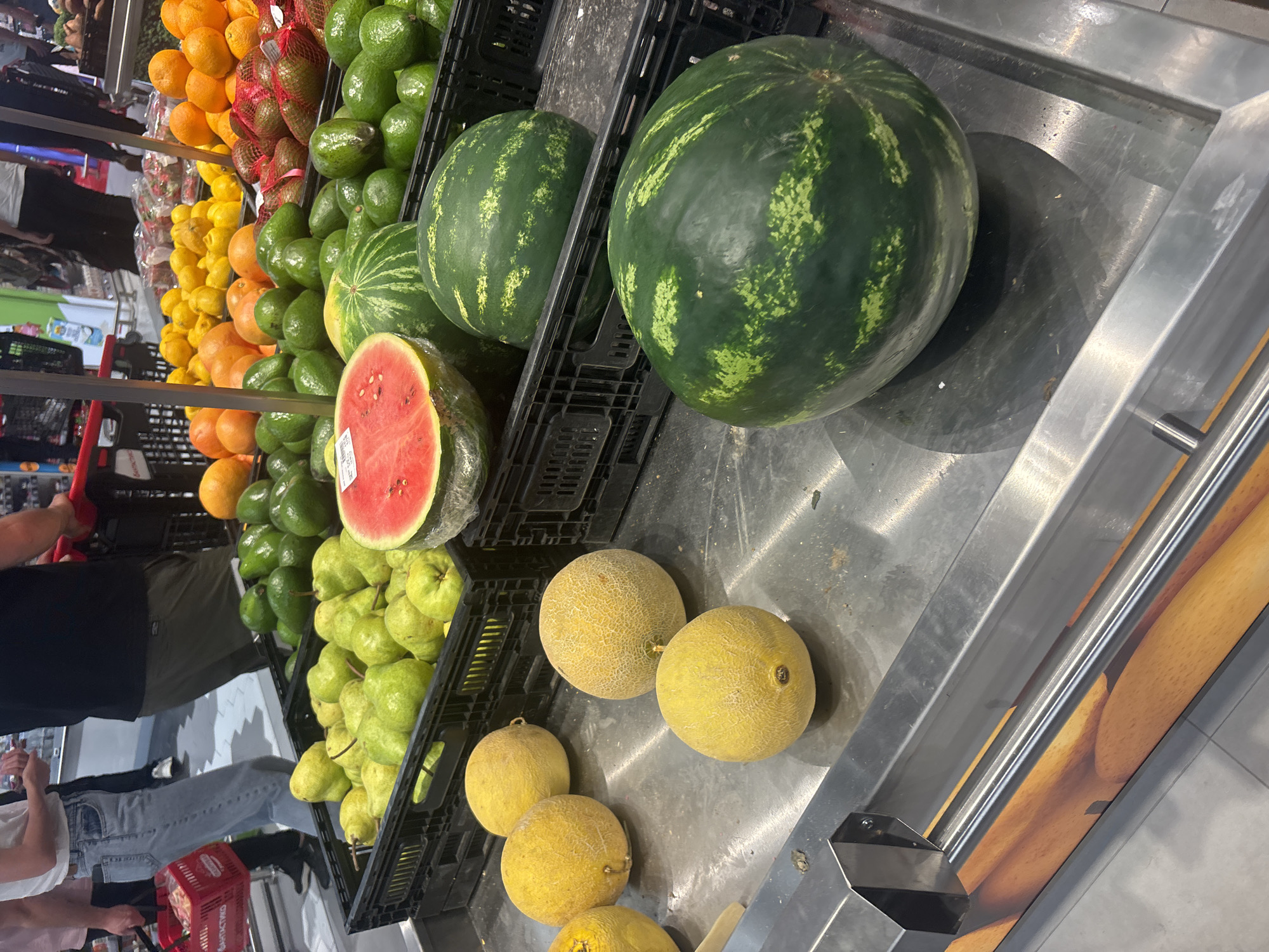 The image depicts a fruit and vegetable display in what appears to be a supermarket setting. Visible fruits and vegetables include oranges, lemons, avocados, watermelons (one sliced open), pears, and cantaloupe. A person wearing jeans and a white shirt is partially visible in the lower-left corner, holding a red shopping basket. The display arrangement includes black plastic crates and a stainless steel surface with wood grain accents on the sides. Given the request for the location in Plovdiv, Bulgaria, it can be inferred that this image was taken inside a store in Plovdiv that stocks the pictured produce. However, the specific store name and address are not identifiable from the visual content alone.