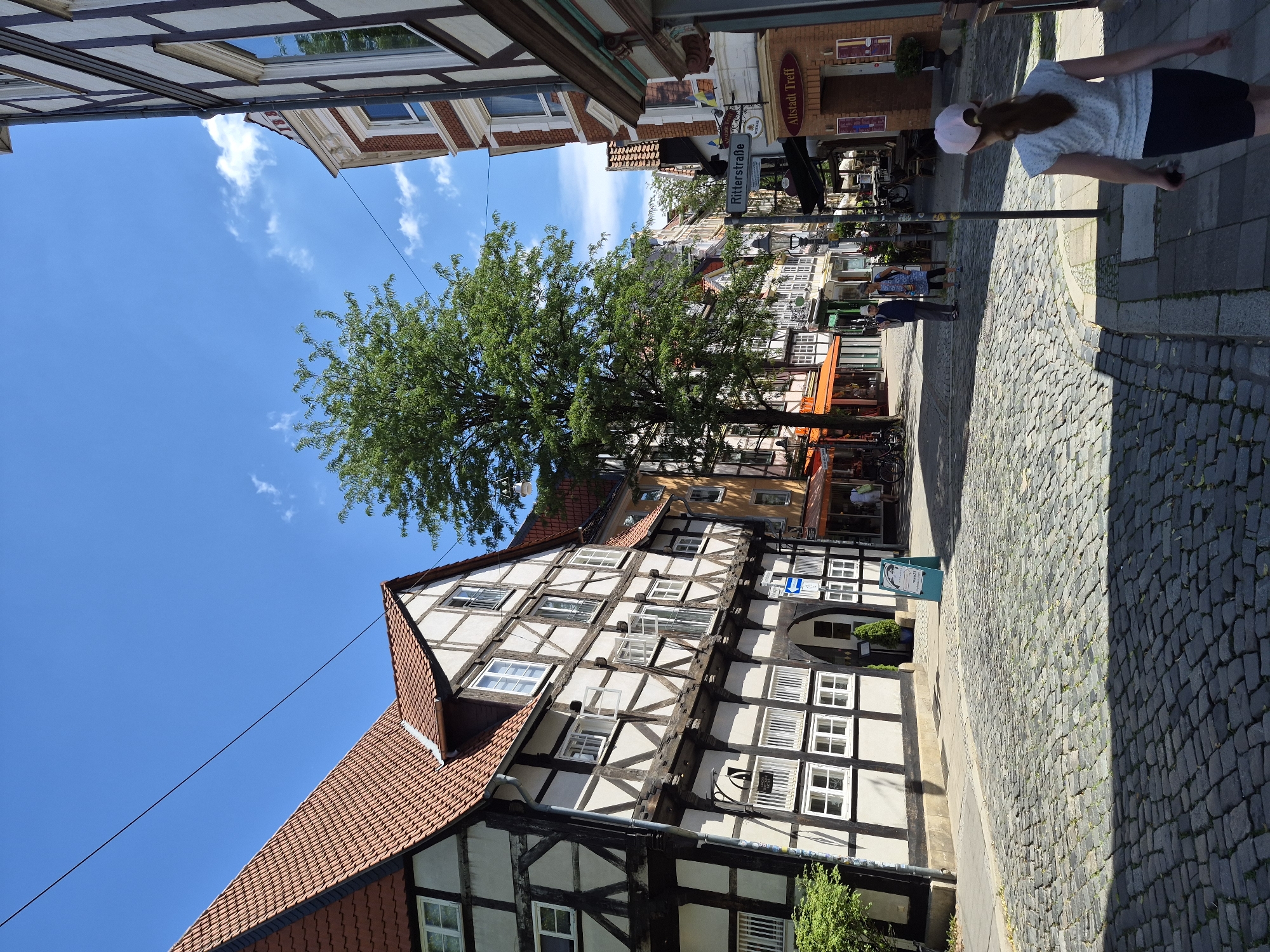 This media file captures a street scene in Brunswick, Germany, under a clear blue sky with scattered white clouds. The photograph is taken vertically, with the buildings and street angled. The architecture features traditional half-timbered houses with red-tiled roofs, characteristic of many German towns.
The scene depicts a pedestrian street paved with cobblestones. On the right side of the street, a woman wearing a light-colored shirt, dark shorts, and a pink cap is walking away from the camera. She is partially in shadow cast by an overhead structure. Further down the street, two other individuals are visible; one in a blue dress and another in a dark top and lighter pants.
In the foreground, a large green tree with dense foliage stands prominently. The buildings lining the street have numerous windows and some display signage. One sign visible on a building to the left reads "Altstadt Treff". Another sign further down the street, also on the left, indicates "Ritterstraße". The presence of awnings over some shopfronts suggests commercial activity. The lighting indicates it is daytime, likely mid-morning to afternoon, given the clear sky and pronounced shadows. The overall impression is a quiet, sunny day in a historic European town.