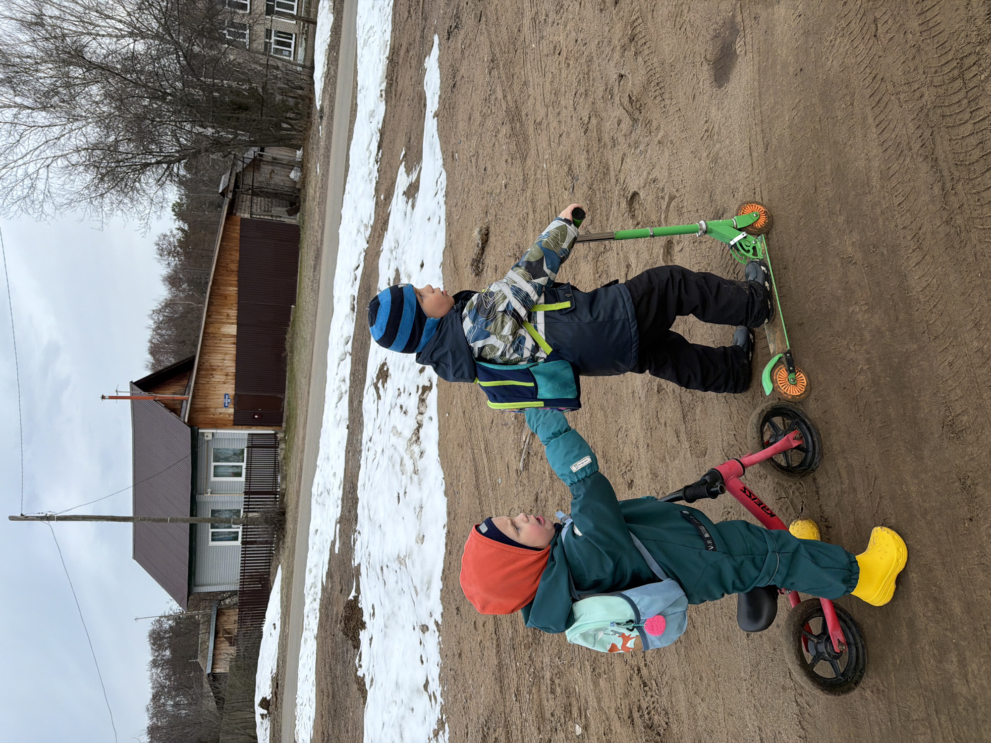 Two young boys are on a dirt and snowy patch of land near a road. One boy is on a green scooter with orange wheels, wearing a blue and black hat, a jacket with a patterned design, and dark pants. The other boy is on a red balance bike with black wheels and is wearing an orange hood, a green jumpsuit, and yellow boots. They are looking at each other and appear to be interacting.
In the background is a residential area with wooden houses and bare trees under an overcast sky. The location is possibly in Chaikovsky, Russia, based on contextual information but no element in the image confirms this.