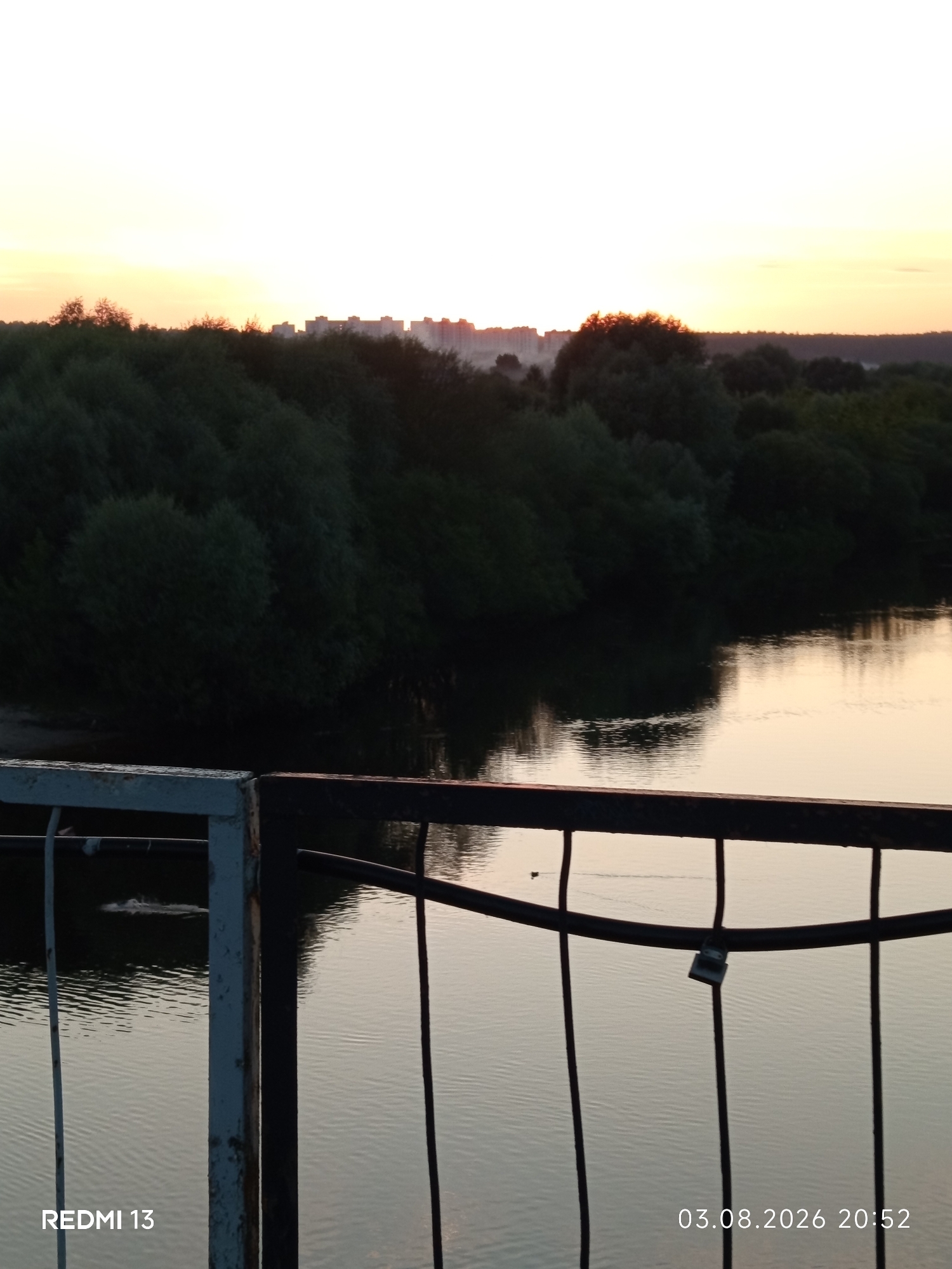 The image captures a tranquil sunset scene over a body of water, likely a river, with a dense line of trees along the opposite bank. In the distance, a cityscape with several multi-story buildings is visible on the horizon, silhouetted against the soft hues of the setting sun. The foreground features a metal railing, partly rusted, with vertical bars and a horizontal bar. A small padlock is attached to the horizontal bar. On the water's surface, ripples are present, and two small ducks are visible swimming. The bottom left corner of the image displays "REDMI 13" and the bottom right corner shows "03.08.2026 20:52," indicating the camera model and the date and time the photograph was taken. The scene is calm and still, suggesting a peaceful evening. This scene is consistent with a view from a bridge overlooking the Berezina River in Barysaw, Belarus, where such urban development and natural landscapes are common.