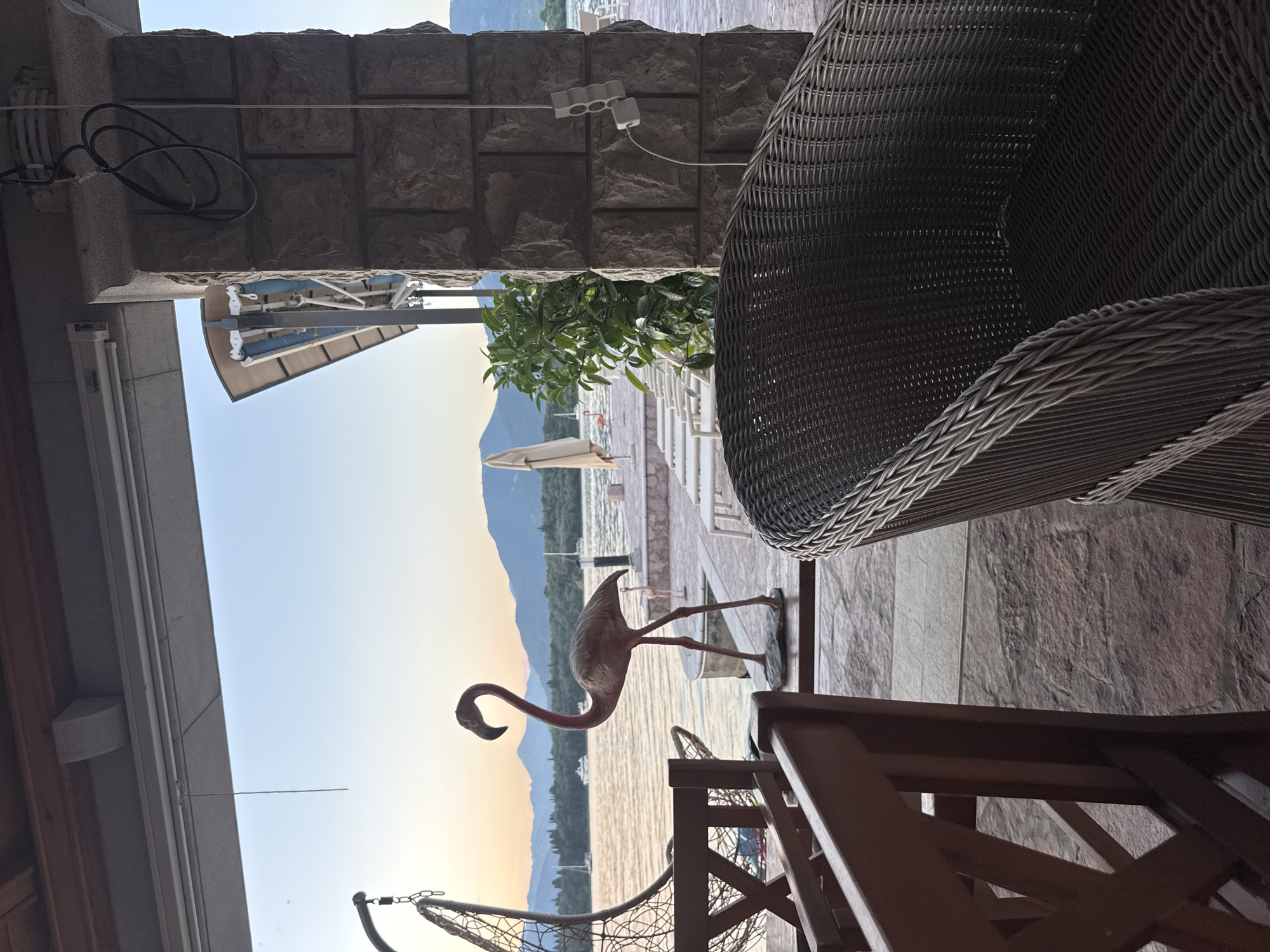 The image depicts a scene in Bogišići, Montenegro. In the foreground, there are elements of a patio or balcony setting, including a wicker chair, a wooden chair frame, and a decorative flamingo statue positioned near the edge. Through the opening, there is a view of the water, a beach area with lounge chairs and an umbrella, and mountains in the background. The lighting suggests either early morning or late afternoon. A stone wall with an electrical outlet is visible above.