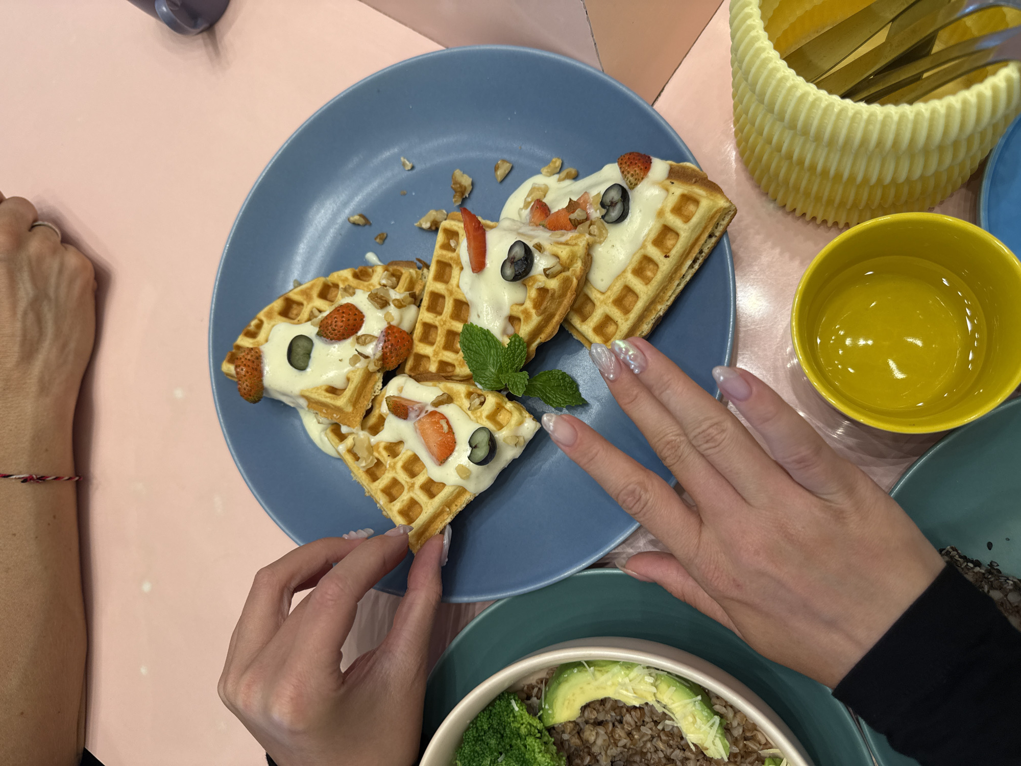 A high-angle shot depicts a meal setting with a variety of dishes on a light pink and off-white patterned surface. A woman's hands are visible, one with a silver ring on the ring finger and a red and white bracelet on her wrist, reaching towards a blue plate containing several pieces of waffles topped with a white sauce, strawberries, blueberries, and chopped nuts. A sprig of mint garnish is placed on top of the waffles. Next to the blue plate is a teal-colored plate holding a bowl with ingredients such as broccoli, sliced avocado, and what appears to be grains. A yellow basket filled with what look like knives or utensils is situated near a small, bright yellow bowl. The woman's other hand is resting near the waffles and the bowl. The scene's location is Sukawati, Indonesia.