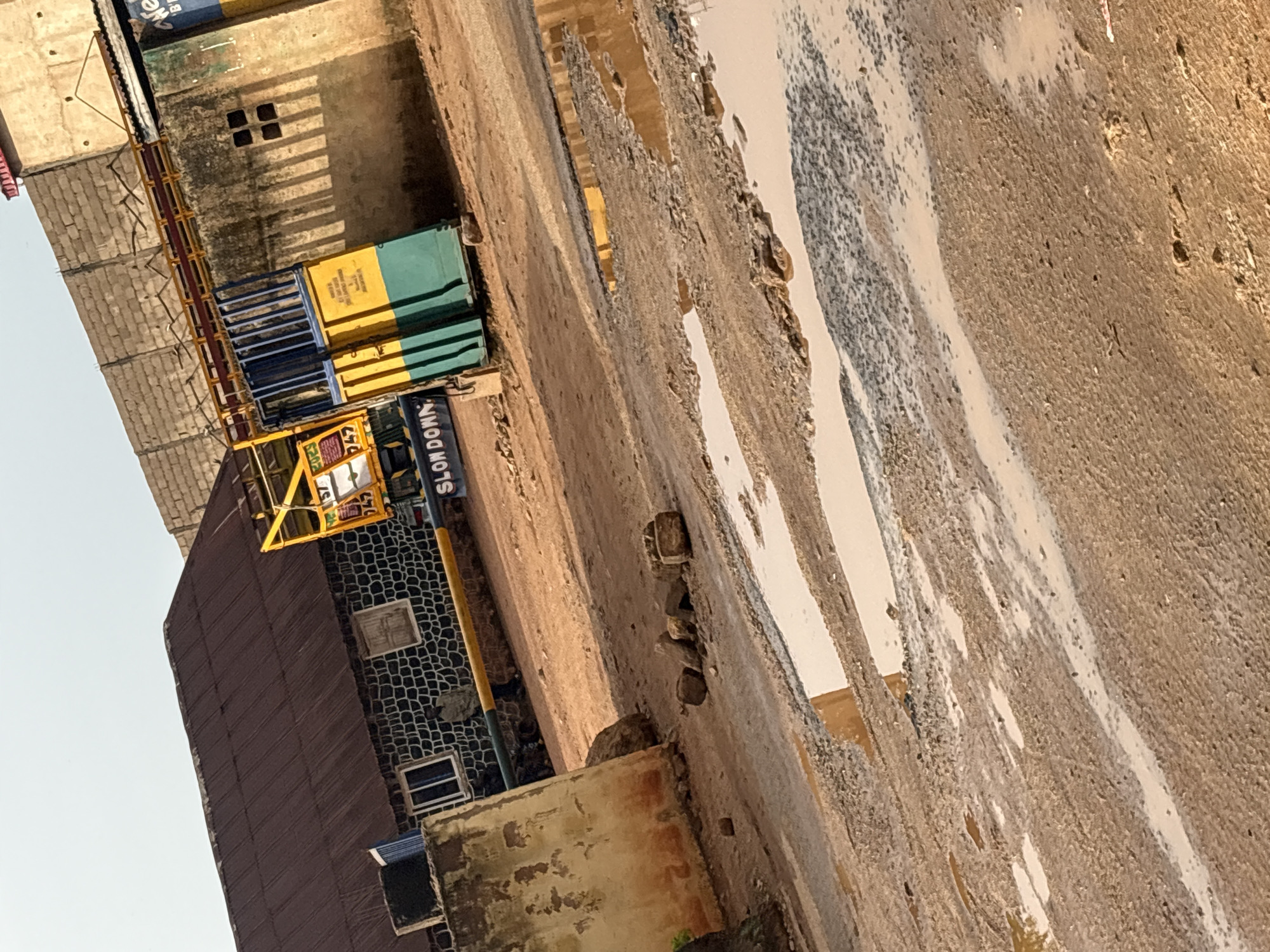 The image depicts a street scene in Bukuru, Nigeria, characterized by visible disrepair of the road surface, featuring potholes filled with water. Buildings line the street, including one with a patterned exterior and another with a "SLOW DOWN" sign. A structure with yellow and blue elements is also present. The image appears to be taken during daylight. There are no people visible in the frame.