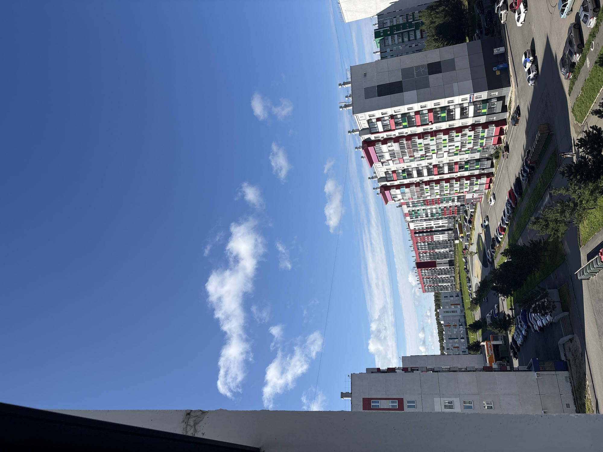 This is an elevated, wide-angle shot of a residential area in Zapadny, Russia, on a clear day with scattered white clouds. In the foreground, on the left, is the side of a white apartment building, partially obscuring the view. The middle ground and background are dominated by several multi-story apartment buildings, painted in various color schemes of white, grey, red, and green. Some buildings feature prominent balconies and window arrangements. Several cars are parked in organized lots and along the streets between the buildings, suggesting a populated urban environment. A sign with Cyrillic text, possibly indicating a store or service, is visible on the side of one building. The presence of trees and a strip of green grass along the roadside suggests some landscaping in the residential area. The overall scene depicts a modern, functional, and well-maintained urban neighborhood.