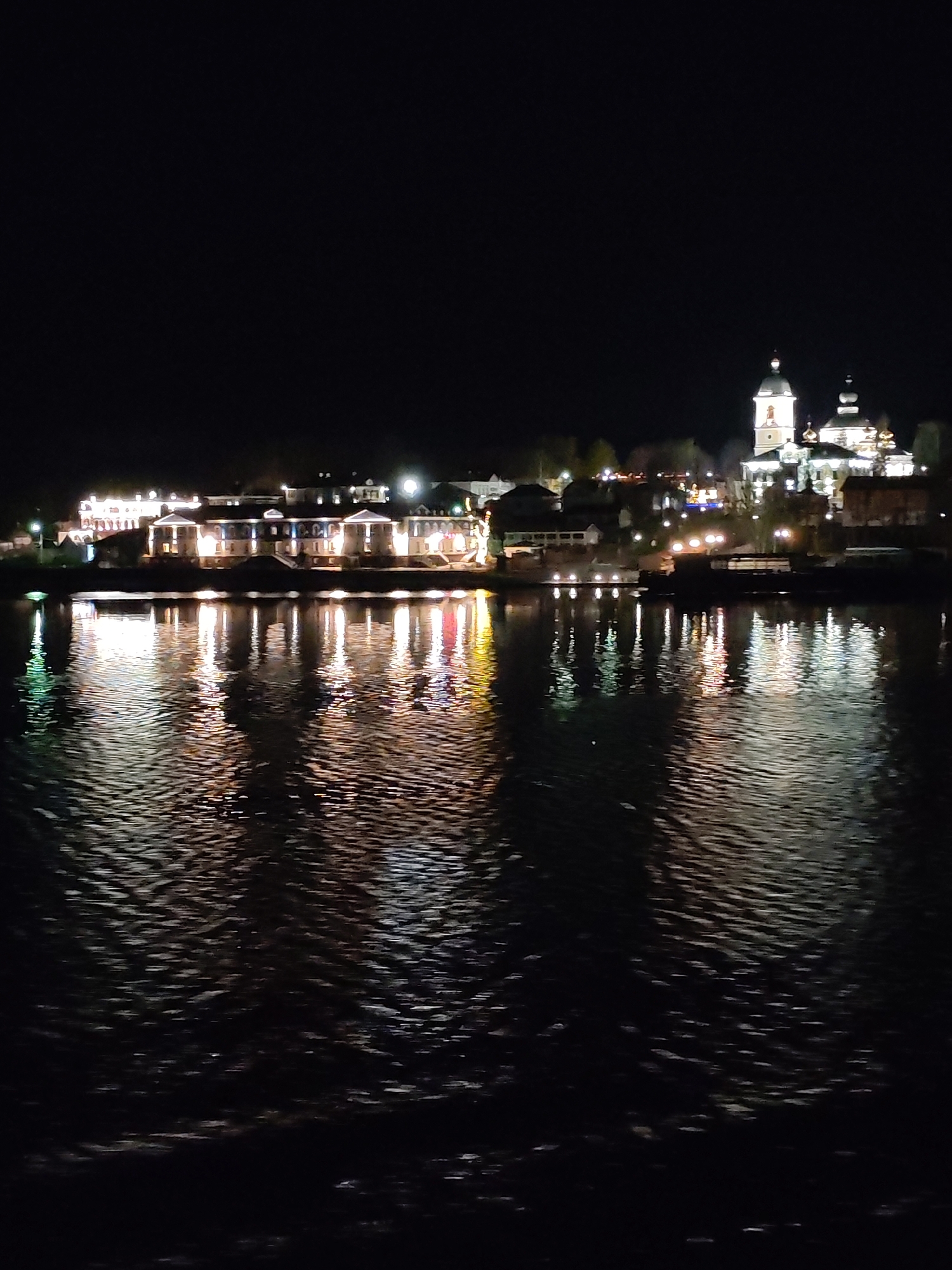 The image depicts a nighttime view of a waterfront in Nesterovo (Slobodskoe S/P), Russia. The foreground shows the surface of a body of water with reflections of lights from buildings on the opposite shore. In the midground are buildings with illuminated windows and architectural features, including a church with distinctive domes. The background is dark, with the night sky. The reflections in the water are distorted by ripples, adding texture to the scene.