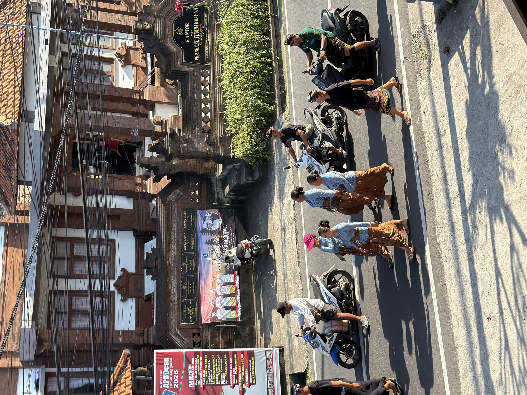 The image shows a procession of people walking on a street in front of buildings with traditional Balinese architecture. The buildings appear to be government offices, as evidenced by the sign that reads "KANTOR PERBEKEL TUMBAKBAYUH" which translates to "Office of the Village Head of Tumbakbayuh". The people are dressed in traditional Balinese attire, with men wearing sarongs and head coverings, and women wearing sarongs and blouses. Some of the people are on motorcycles, while others are walking. A large banner is displayed on one of the buildings, which appears to be an informational poster related to village finances, titled "INFOGRAFIK APBDesa Tahun Anggaran 2026". The scene is illuminated by bright sunlight, casting long shadows on the road. The environment suggests a community gathering or event taking place in the village of Tumbakbayuh, Mengwi, Indonesia.