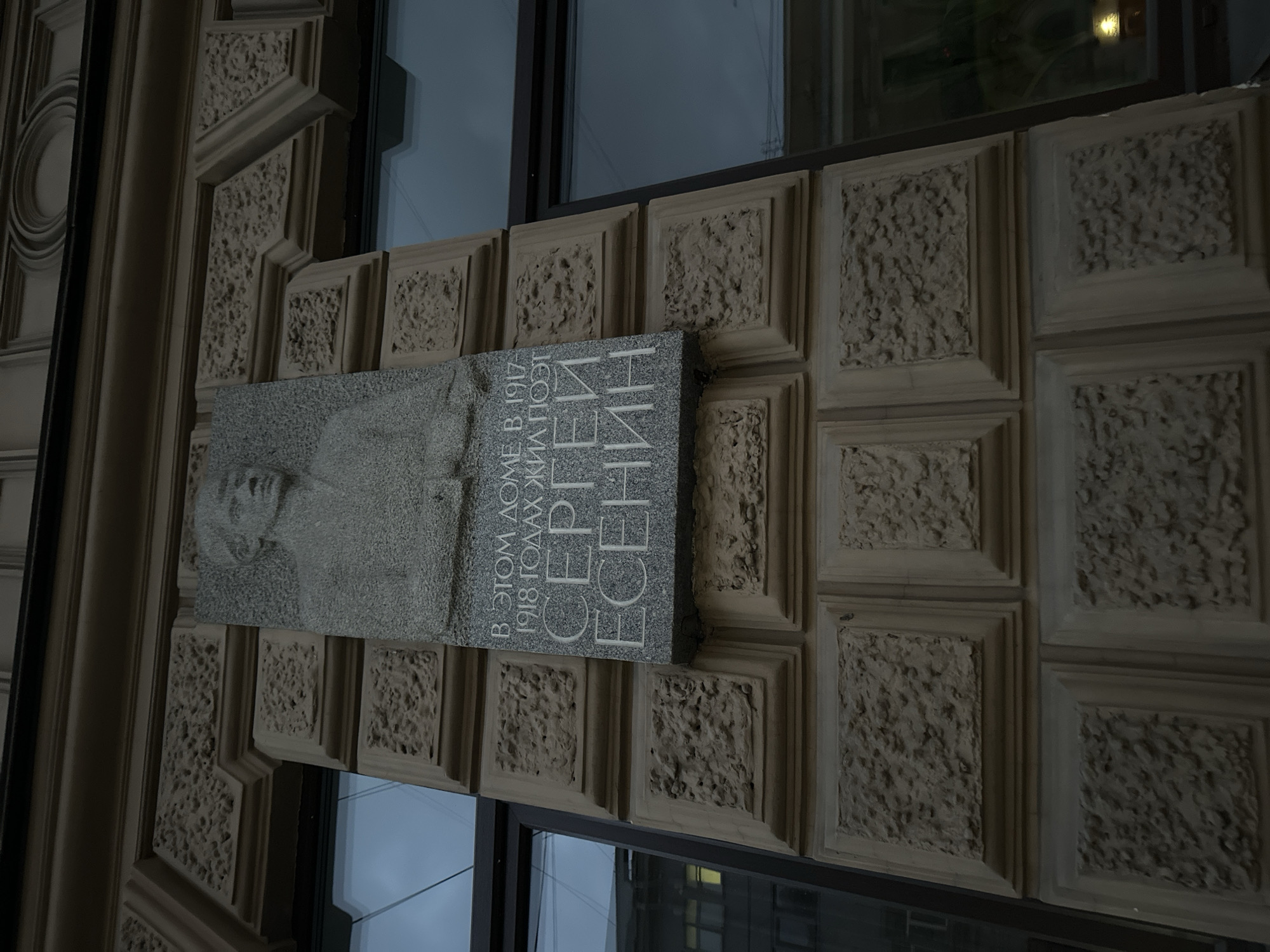 The image captures a memorial plaque in Saint Petersburg, Russia, dedicated to the poet Sergei Yesenin. The plaque is mounted on the exterior of a building with heavily textured facade and window fixtures. The plaque features a relief portrait of Yesenin and Cyrillic text, identifying him and noting his residence in the building between 1917 and 1918. The lighting is subdued, with details appearing muted due to overcast conditions or low light. The exterior architecture is indicative of a classical or neoclassical style, marked by its ornamental facade.