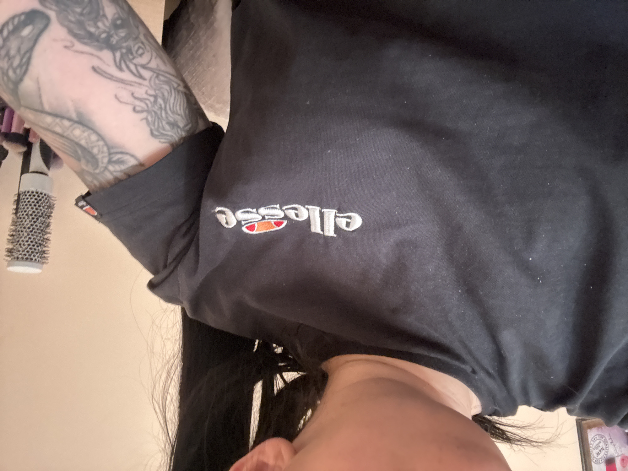 The image depicts a person wearing a black t-shirt with the "ellesse" logo embroidered on the front. The person's left arm is visible, covered in a detailed tattoo. Part of the person's face, including an ear and part of the chin, is also in the frame. A hairbrush is seen in the background, hanging on a wall. The image appears to be taken indoors, possibly in a bedroom or bathroom. There is no information available in the image to confirm the location of Plovdiv, Bulgaria.