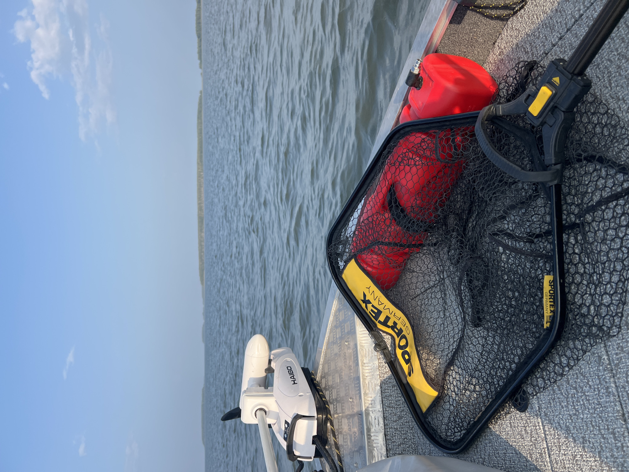 An eye-level, medium shot captures a fishing net, a red jerrycan, and a white electric trolling motor on a boat deck. The boat is on a large body of water under a pale blue sky with scattered white clouds. The horizon is lined with distant islands covered in trees. The fishing net, with a black frame and mesh, has a yellow tag with "SPORTEX GERMANY" printed on it. Inside the net, a red flotation device is visible. The jerrycan is also red and appears to be made of plastic. The trolling motor, mounted on the edge of the boat, is white with the brand name "HAIBO" visible. The deck of the boat is made of gray material with a metallic undertone. The scene is set in Novopichugovo, Russia.