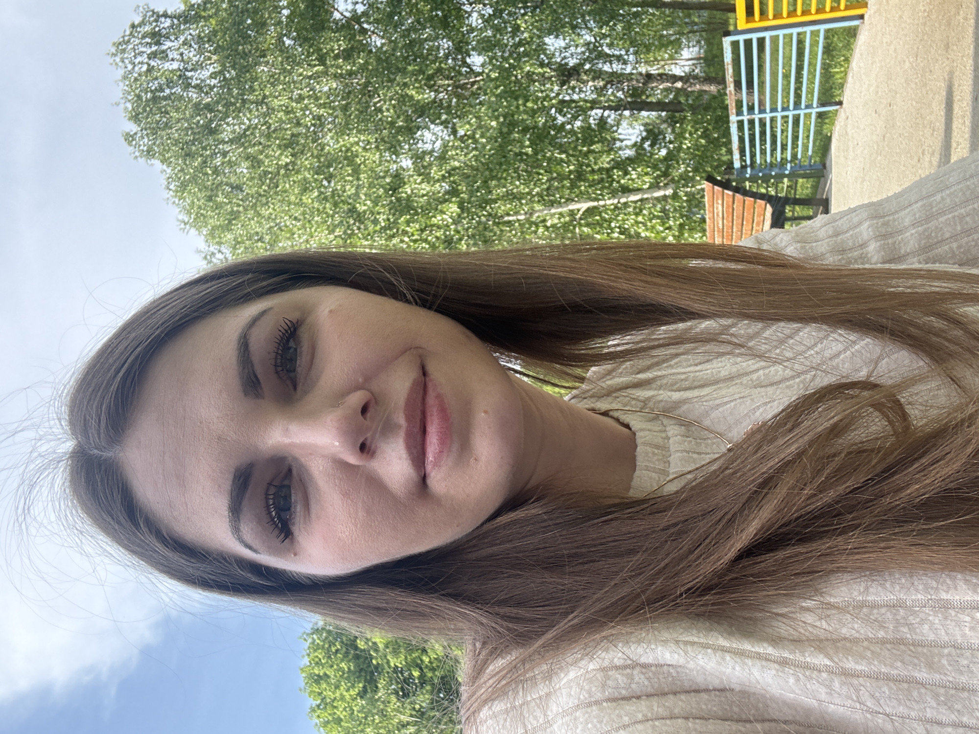 A young woman is pictured from a close, slightly upward angle, potentially in Shevlyakovo, Russia (though no specific details confirm this). Her long brown hair is visible, and she is wearing a light-colored, textured sweater. Behind her, green foliage and trees are visible, along with a partially visible playground structure with yellow and blue bars and a brown bench. The sky is visible in parts of the background. The woman's face is in focus, showing her facial features, including her eyes, nose, and lips.