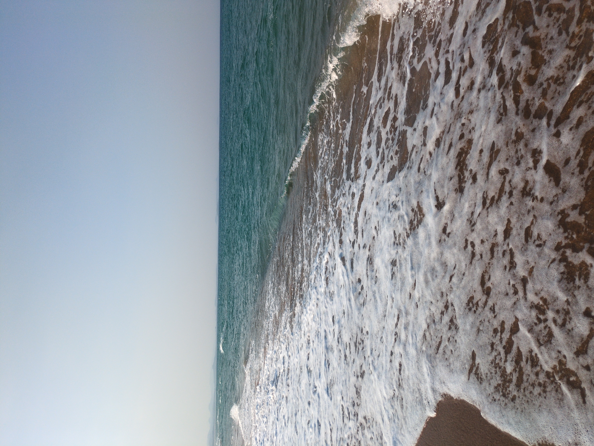 A sideways view shows a beach scene. On the left is a light blue sky. The water on the beach is a deeper blue-green color. White foam from the waves covers a portion of the water and the beach on the right. The location is Tuy Hoa, Vietnam.