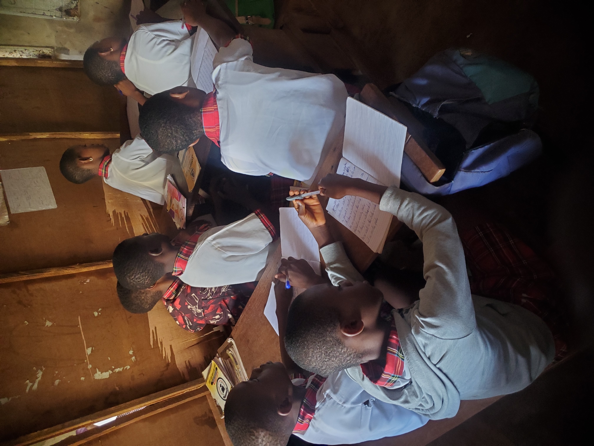 The image shows seven schoolchildren sitting at wooden desks in a classroom. All are dressed in white shirts with red plaid collars. They are writing in notebooks with pens. Some have books stacked on their desks. A blue backpack is also on one of the desks. The classroom appears to have wooden walls and floors. The location is Unknown, Unknown.