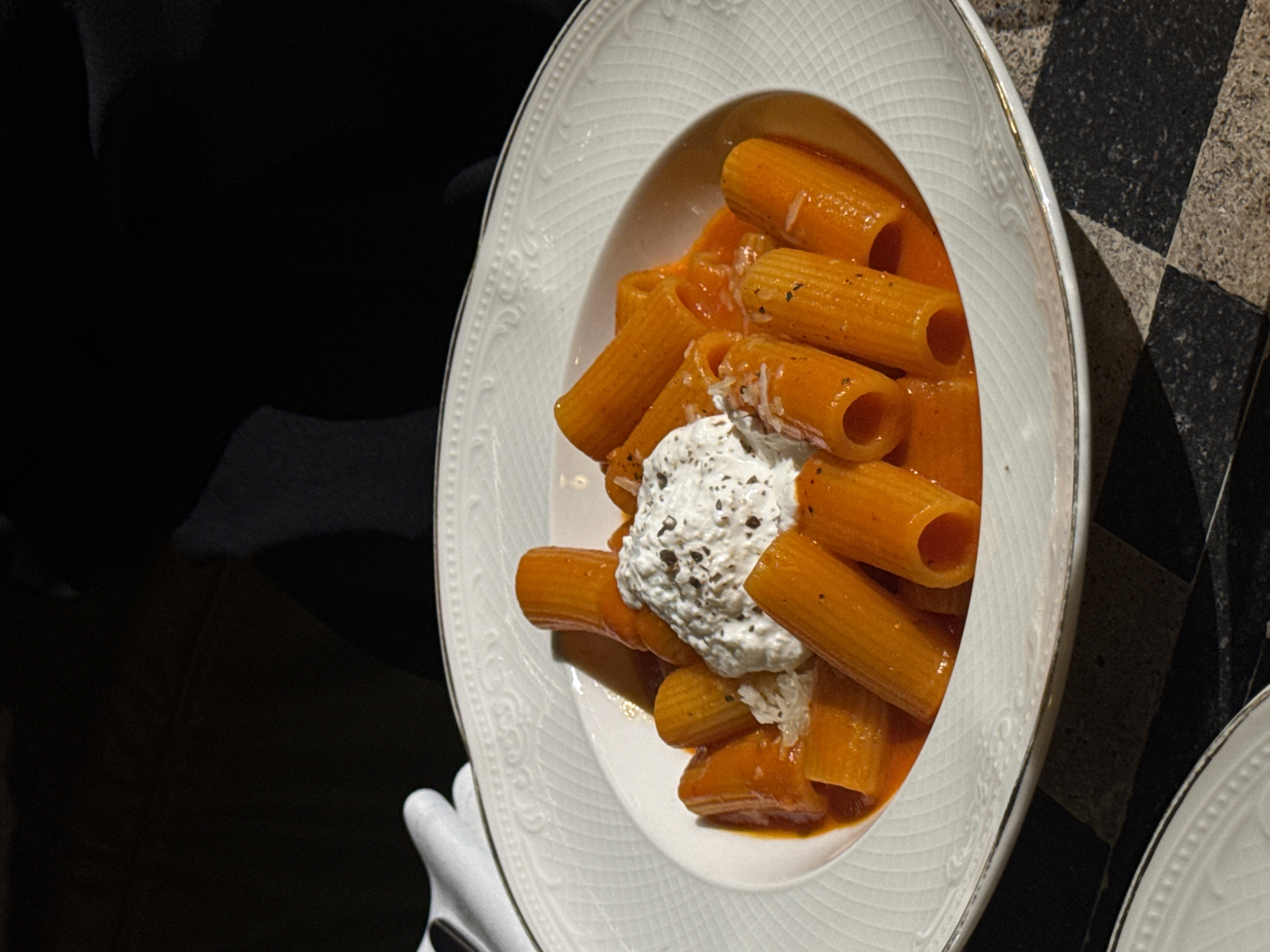 A high-angle shot depicts a white, ornate oval plate filled with rigatoni pasta covered in tomato sauce and topped with a dollop of what appears to be ricotta cheese and herbs. The plate is positioned on a surface with a black and white checkerboard pattern. A person wearing a dark-colored garment is holding the plate, as evidenced by a gloved hand visible in the lower-left corner. A portion of another similar plate is visible in the lower-right corner. The location is likely a restaurant in Yerevan, Armenia.