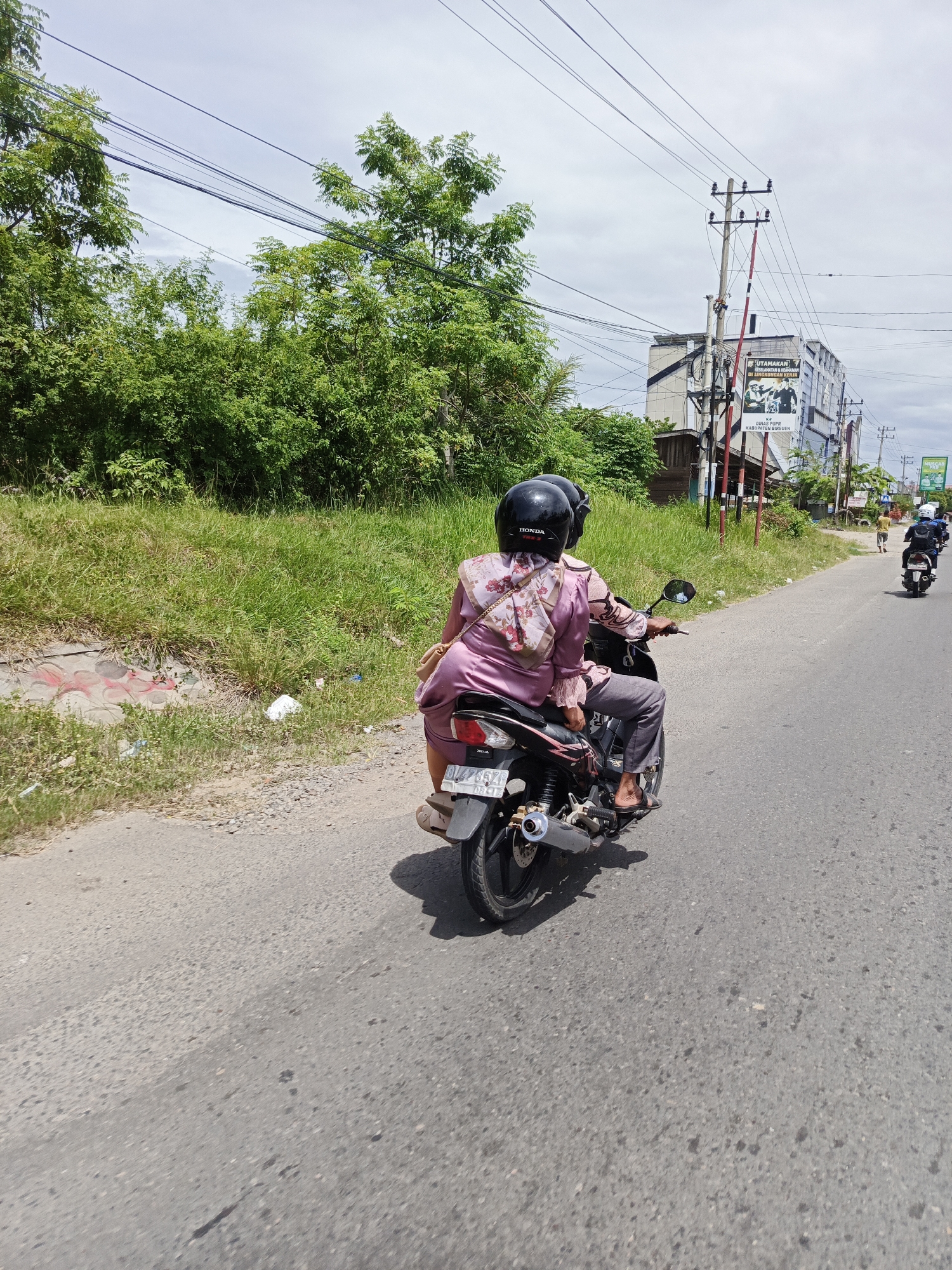 This is a daylight, eye-level, medium shot of two individuals riding a motorcycle on a paved road in Cotbadatunong, Indonesia. The rider and passenger both wear black helmets. The passenger is in front, wearing a light pink blouse with a floral pattern and grey pants. A light brown shoulder bag is slung over her left shoulder. The motorcycle appears to be a dark color. The road is bordered by a grassy embankment on the left and a line of trees. In the background, buildings and utility poles are visible, with a large banner on one of the buildings. There are other motorcycles in the distance on the road. The sky is cloudy.