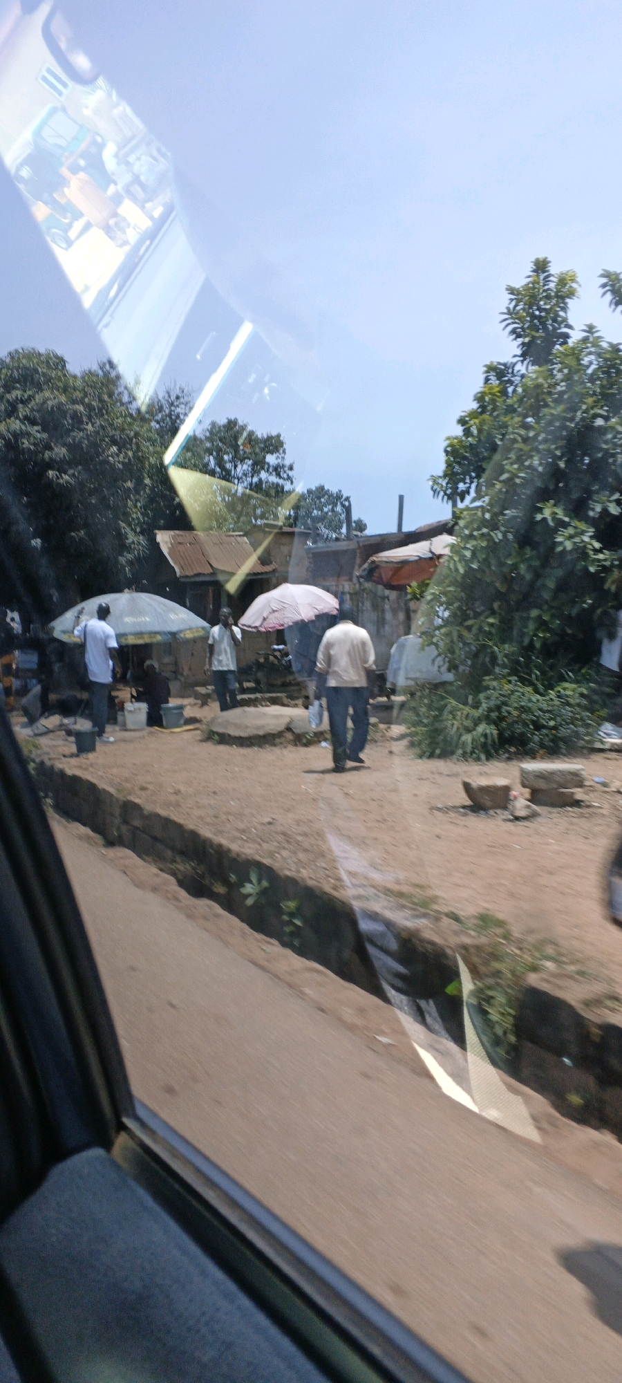 A wide shot captures a street scene with several individuals and market stalls. The foreground features a dusty road with a narrow drainage ditch. In the midground, a few people are gathered around market umbrellas, with one person in the center walking away from the camera. Buildings with corrugated metal roofs are visible in the background, alongside dense foliage. The sky is clear and blue. The image appears to be taken from inside a vehicle, with reflections from the vehicle's interior visible on the surface of the glass.