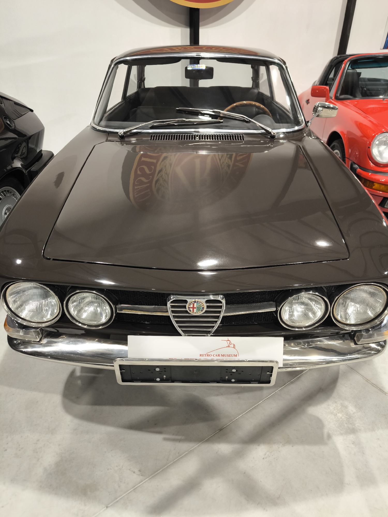 The image shows the front of a brown Alfa Romeo parked in a showroom. A white sign with the text "RETRO CAR MUSEUM" in red lettering partially obscures the front bumper. The car has four round headlights and a chrome bumper. A red sports car is partially visible in the background on the right side of the image. A black car is partially visible in the background on the left side of the image. The showroom has a white floor and walls. There is a sign in the background that is partially visible. The city of Kapatovo, Bulgaria is not discernible from the image.