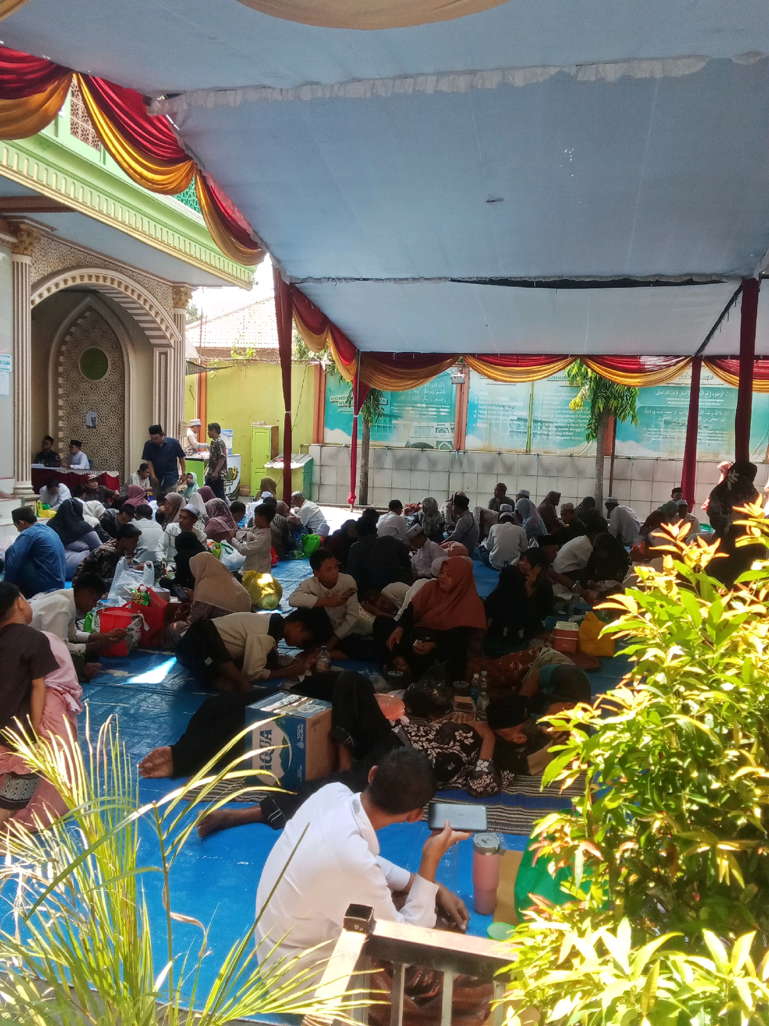 The image depicts an outdoor gathering under a large, light blue tent with red and gold draped fabric at the edges. A green and white building with intricate architectural details, likely a mosque, is visible on the left. Numerous people, dressed in traditional Islamic attire, are seated on a blue mat, appearing to be engaged in a religious or community event. Many are wearing head coverings, including hijabs and caps. Some individuals are interacting, while others appear to be listening or contemplating. Objects scattered on the mat include bags, a cardboard box with "AQUA" branding, and water bottles. In the foreground, lush green foliage is prominent, suggesting an outdoor setting. The scene appears to be located in Jabontegal, Indonesia, given the architectural style and the presence of what seems to be a mosque. The overall atmosphere is one of quiet congregation and communal gathering.