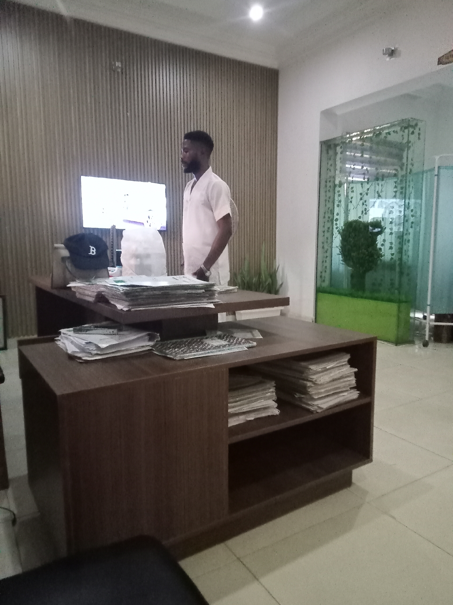 The image depicts an office interior in Gwarinpa, Nigeria. A man with a beard and short hair, wearing a white shirt, is standing next to a desk. The desk is made of dark brown wood and has two levels. The top level is cluttered with papers, a black baseball cap with the letter "B" on it, and a white plastic bag. The bottom level contains two shelves filled with stacks of papers. Behind the desk is a television screen, and a wall with vertical wooden panels. To the right is a glass enclosure containing a green plant. The floor is tiled. A black chair is partially visible in the foreground.