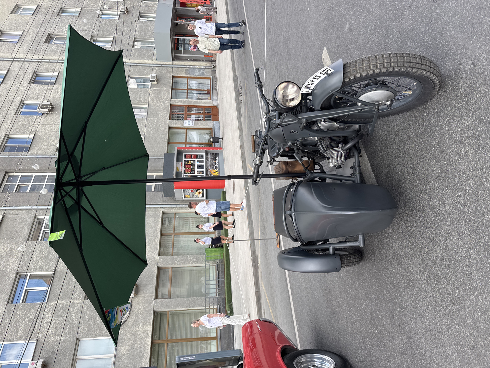 The image depicts a Zündapp KS 600 motorcycle with a sidecar parked on a paved street in Novosibirsk, Russia. A large green umbrella is mounted over the motorcycle. In the background, a multi-story building with numerous windows and air conditioning units lines the street. Several people are visible on the sidewalk: a woman in white, three individuals in uniform, and two men walking. A red car is partially visible to the left. A kiosk with "GRILL" signage is also present. The overall scene suggests an outdoor event or exhibition.