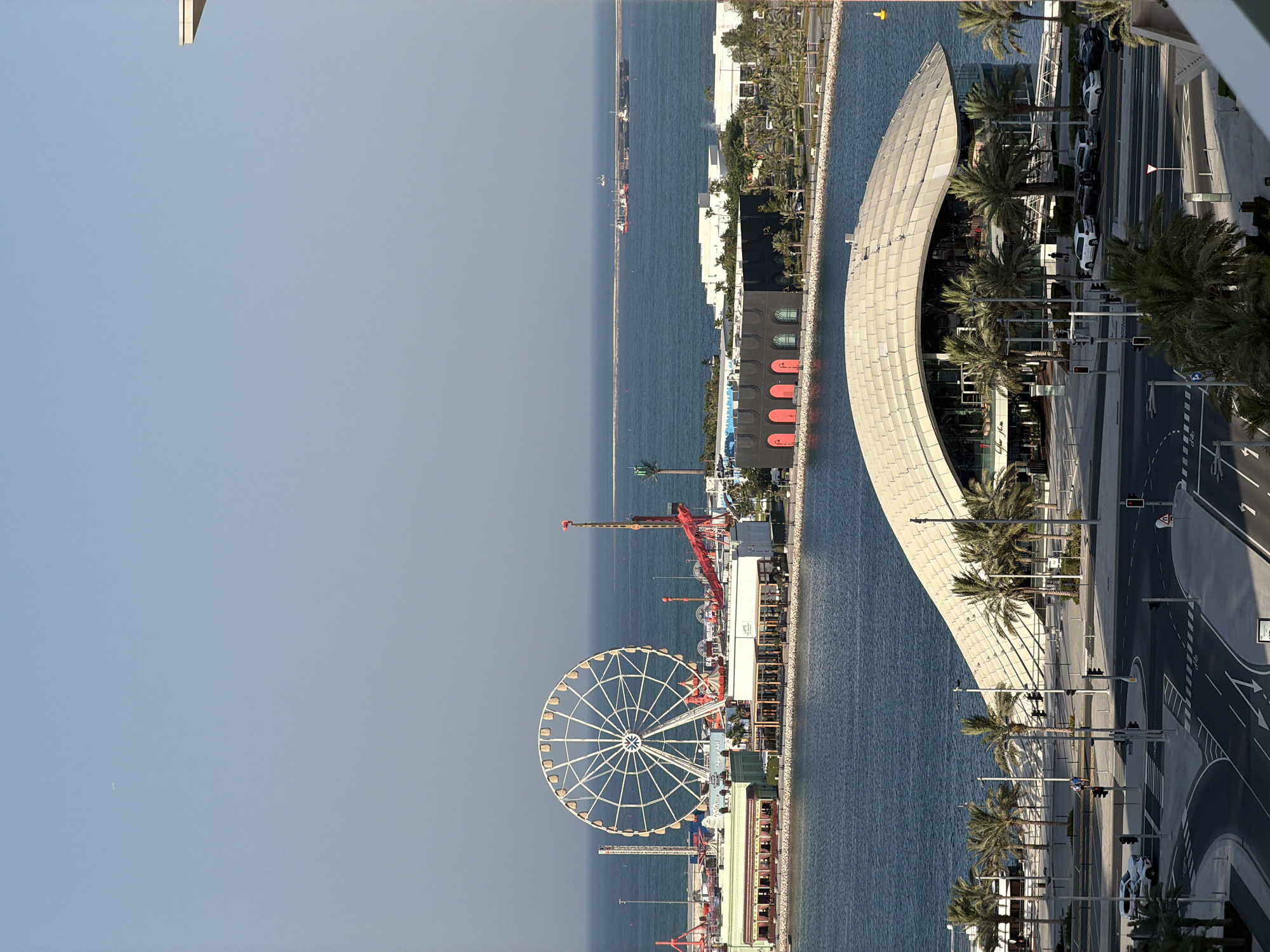 This is a high-angle, vertical-orientation, outdoor shot of a waterfront promenade. The scene is bathed in bright sunlight, suggesting it is daytime, likely late morning or afternoon, with clear skies. The water is a deep blue, reflecting the sunlight.
The foreground on the right features a modern, curved building with a pale, textured facade. In front of the building, a paved road with traffic lights and lane markings is visible, with several white cars parked or driving. A row of palm trees lines the street, casting shadows on the road and sidewalk.
Further into the scene, along the waterfront, a large Ferris wheel stands prominently, suggesting an amusement area or park. There are also cranes and other structures associated with a port or entertainment district. Ships are visible in the distance on the water, and a breakwater or pier extends into the sea.
Along the promenade, there are buildings with distinctive architecture, including some with red arched windows. The overall setting appears to be a developed urban area with leisure and commercial activities, possibly a tourist destination. No people are clearly discernible in the frame, but the presence of cars and the general infrastructure suggest human activity. There is no discernible text in the image.