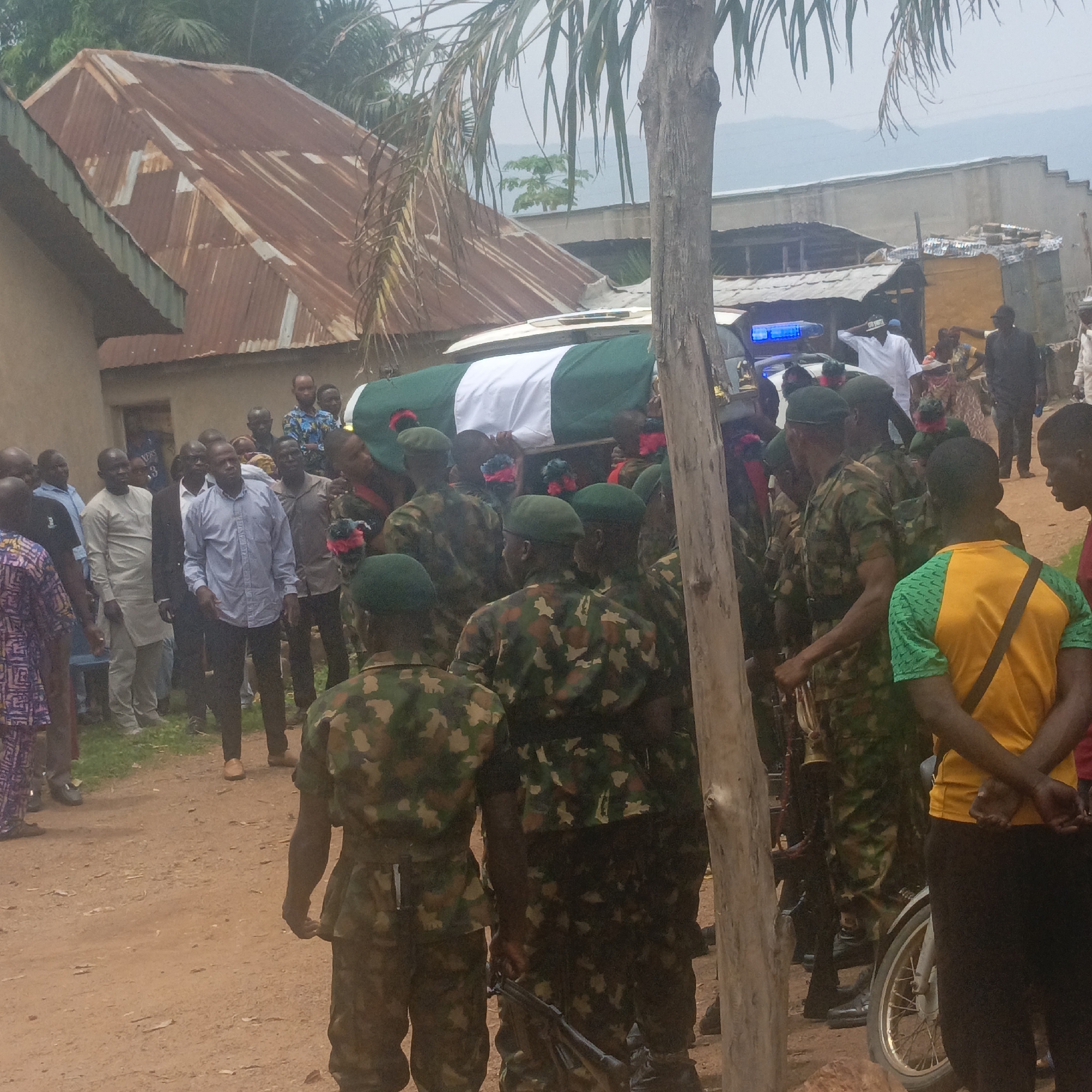 The image depicts a funeral procession in what appears to be a rural area in or near Akwanga, Nigeria. A coffin draped with the Nigerian flag is being carried by soldiers in camouflage uniforms and green berets. Several people, including civilians dressed in various attires, line the sides of the dirt road, watching the procession. In the background, there are houses with corrugated iron roofs and other structures. There is also a motorcycle parked nearby. The overall environment suggests a solemn and respectful atmosphere. The presence of armed soldiers implies it is a military burial.