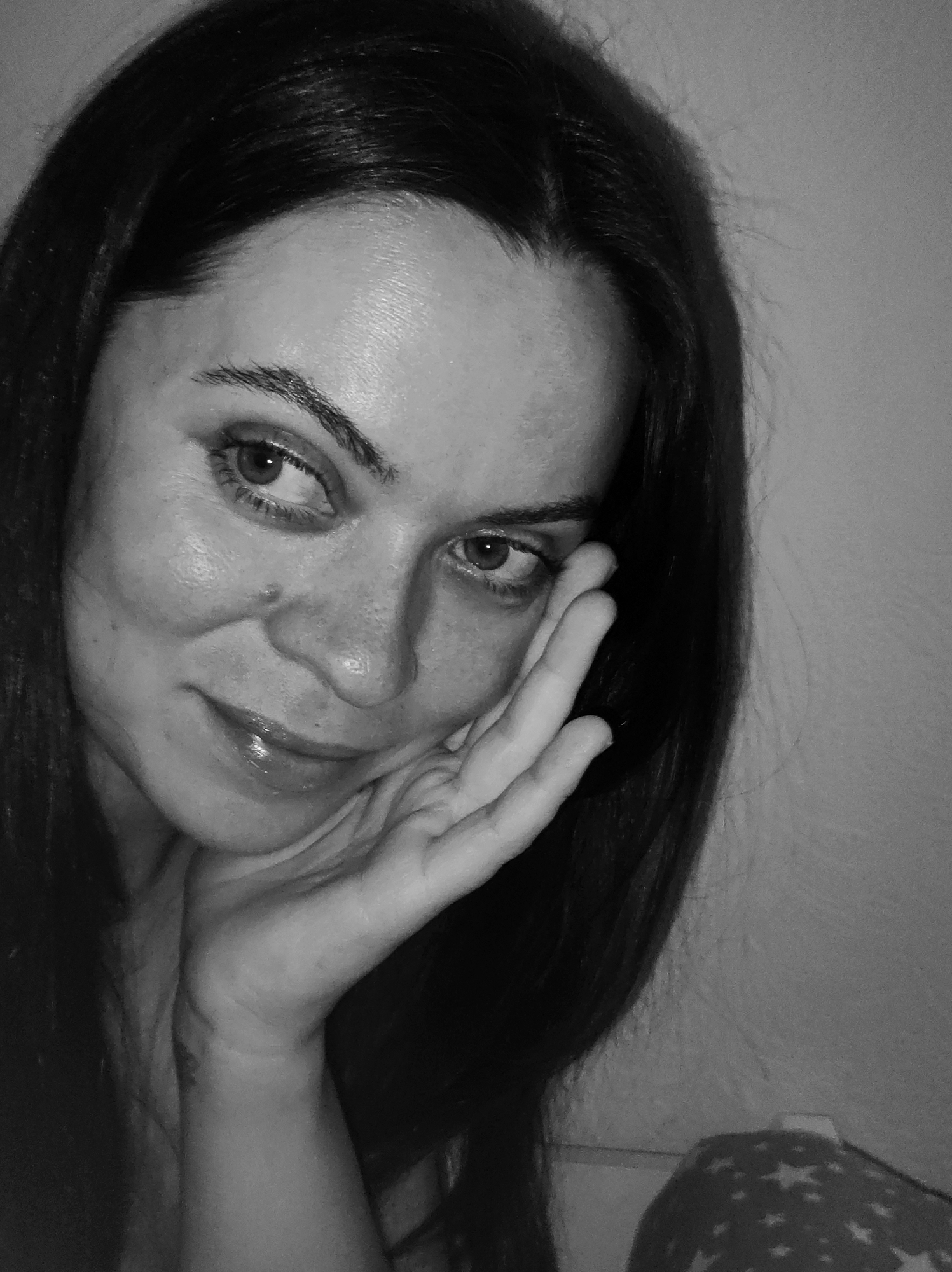 A black and white close-up portrait shows a woman with long, dark hair resting her face on her left hand. She is looking off to the side with a slight smile. The background appears to be a plain wall on the right and a patterned fabric or clothing is visible at the bottom right corner. The image gives no indication of location in the city of Chaikovsky, Russia.
