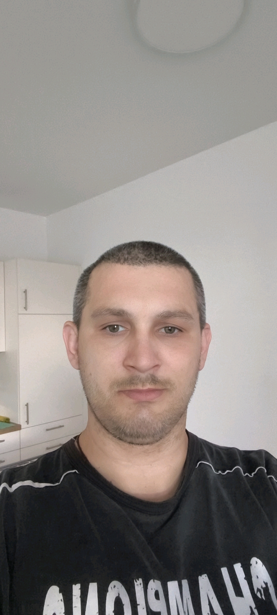 A man with short, greyish-brown hair and light brown eyes is looking directly at the camera. He has a light stubble and a faint smile. He is wearing a black t-shirt with a white design across the chest. He is standing in what appears to be a kitchen, with white cabinets visible in the background. The overall lighting is bright and even. The image does not provide enough information to determine the specific location in Wolgast, Germany.