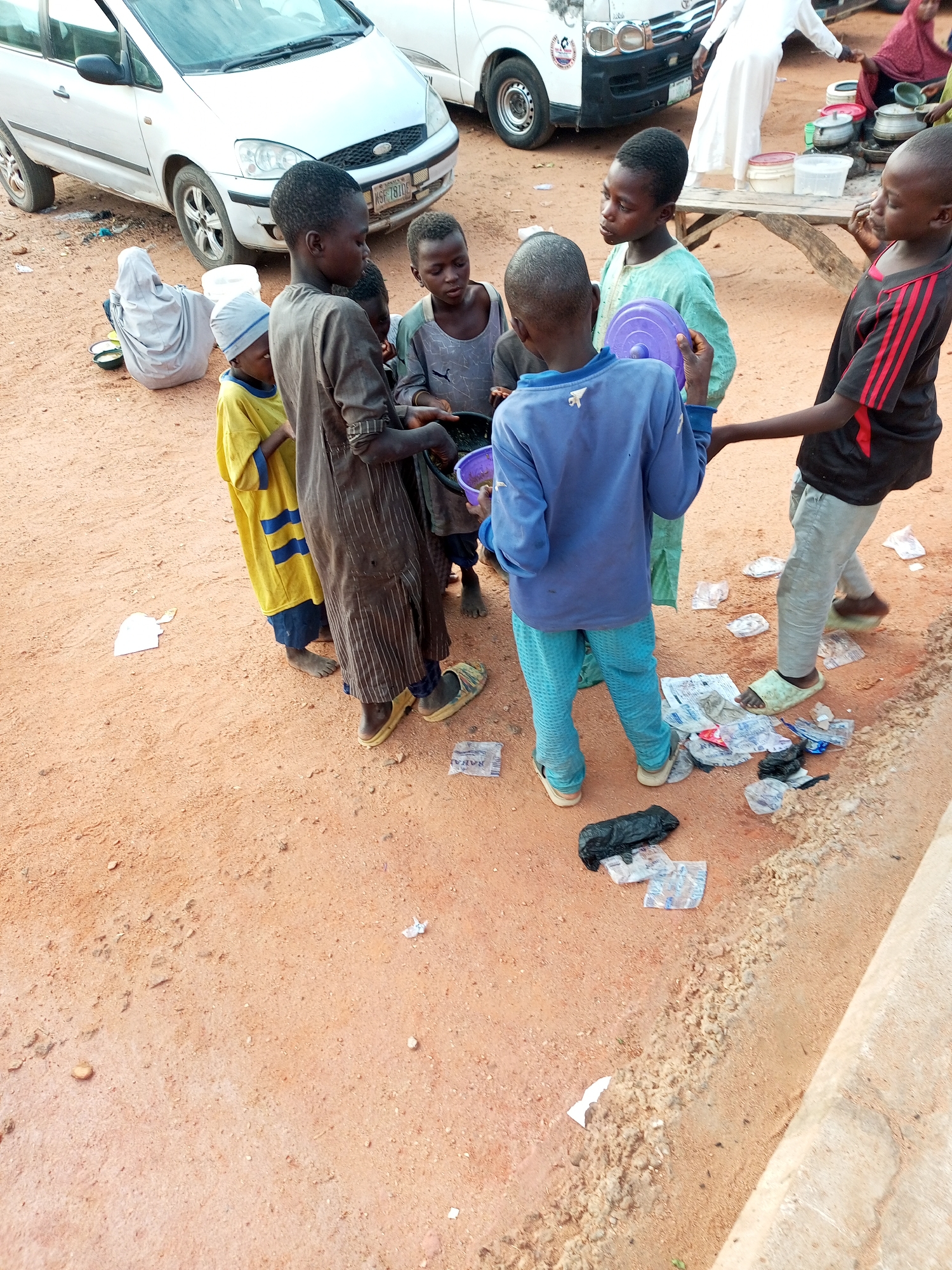 The image shows a group of children gathered on a dirt surface next to a white vehicle. One child, wearing a blue shirt and light blue patterned pants, is holding a purple lid. Another child in a brown garment with vertical stripes is also holding a purple lid. A child in a yellow shirt with blue stripes stands nearby, and another child in a green shirt is also present. To the left, a person is seated on the ground, wearing a light gray hooded garment, with several shallow containers in front of them. In the background, another vehicle and a wooden table with cooking pots are visible. The ground is covered with dirt, scattered plastic wrappers, and a black plastic bag. The scene appears to be outdoors.