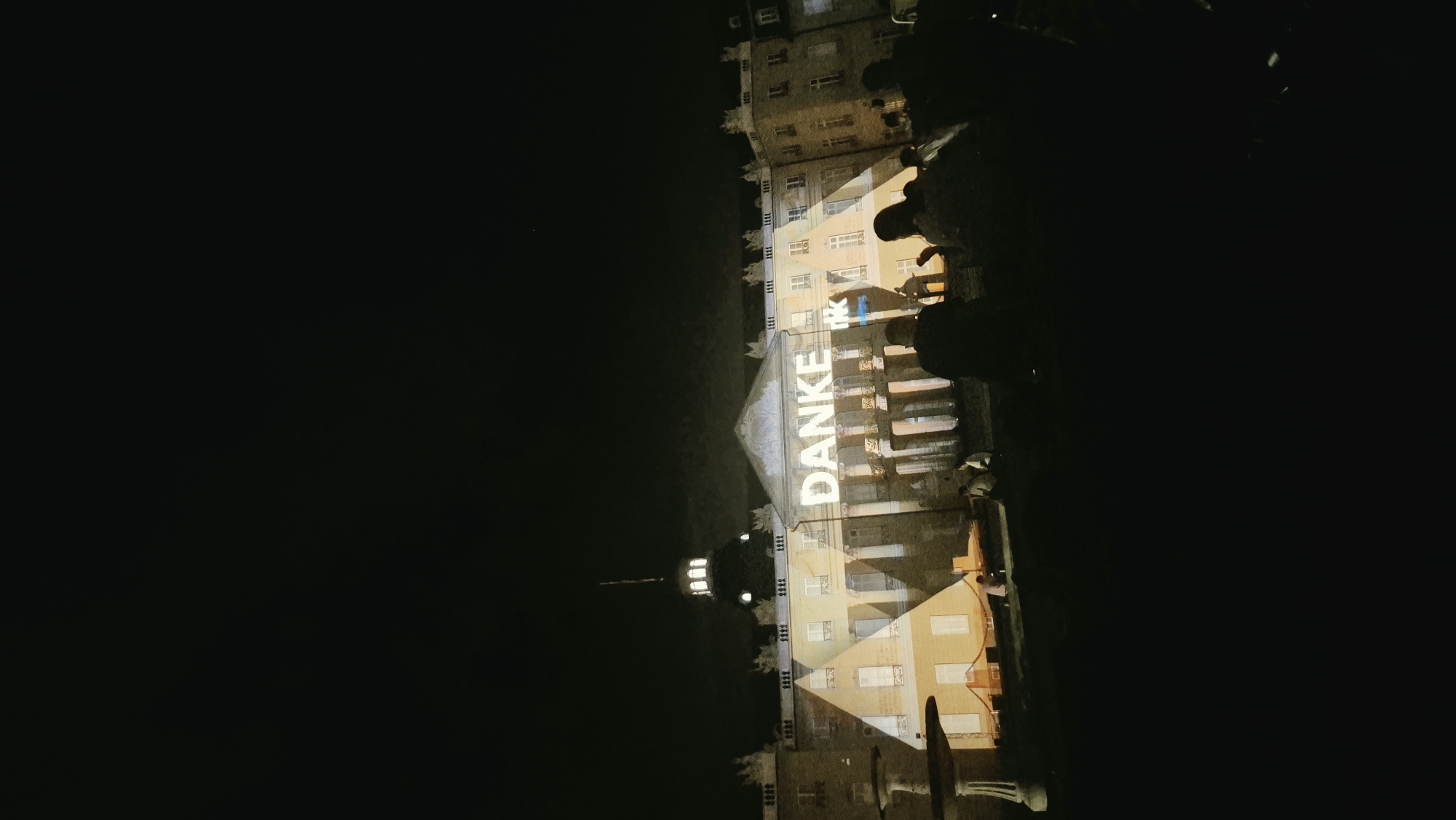 In Karlsruhe, Germany, at night, a large building, likely a palace or historic structure, is illuminated by a projection of the word "DANKE," which means "Thank you" in German. The word is projected across the facade of the building, with beams of light extending outwards. A crowd of people are visible in silhouette in the foreground, facing the building, suggesting they are gathered to watch the projection. The environment is dark, indicating it is nighttime. The architecture of the building appears classical, with multiple windows and a prominent central section.