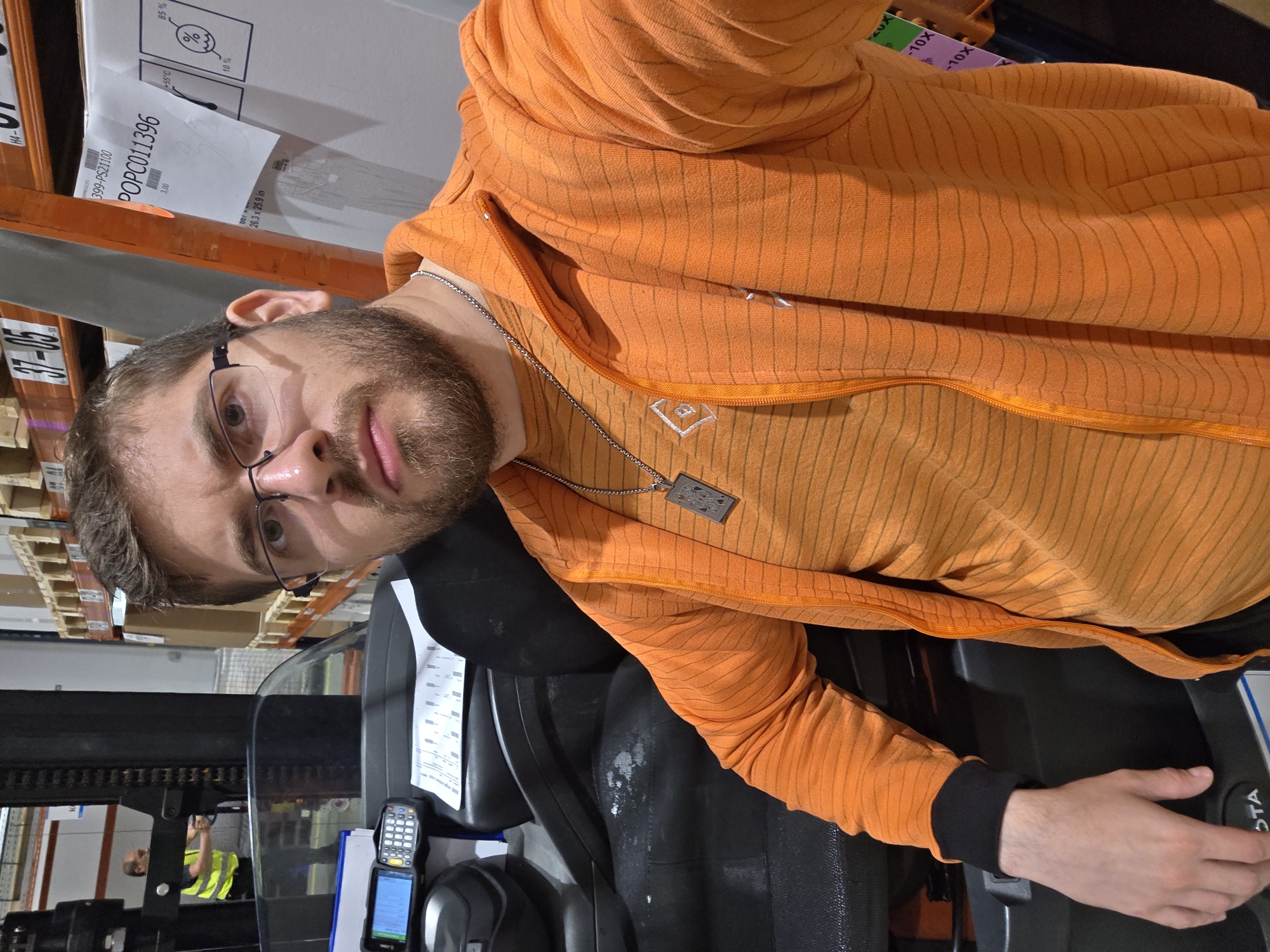 The image shows a man with glasses and a beard, wearing an orange jacket and a necklace, sitting in a forklift. A handheld barcode scanner and a clipboard are visible near the forklift controls. Pallet racking with boxes is visible in the background. Text visible on one of the boxes includes "399-PS21100" and "POPC011396." A man wearing a yellow safety vest is visible outside the forklift. The location is likely a warehouse or distribution center in Plovdiv, Bulgaria, but this cannot be conclusively determined from the image alone.
