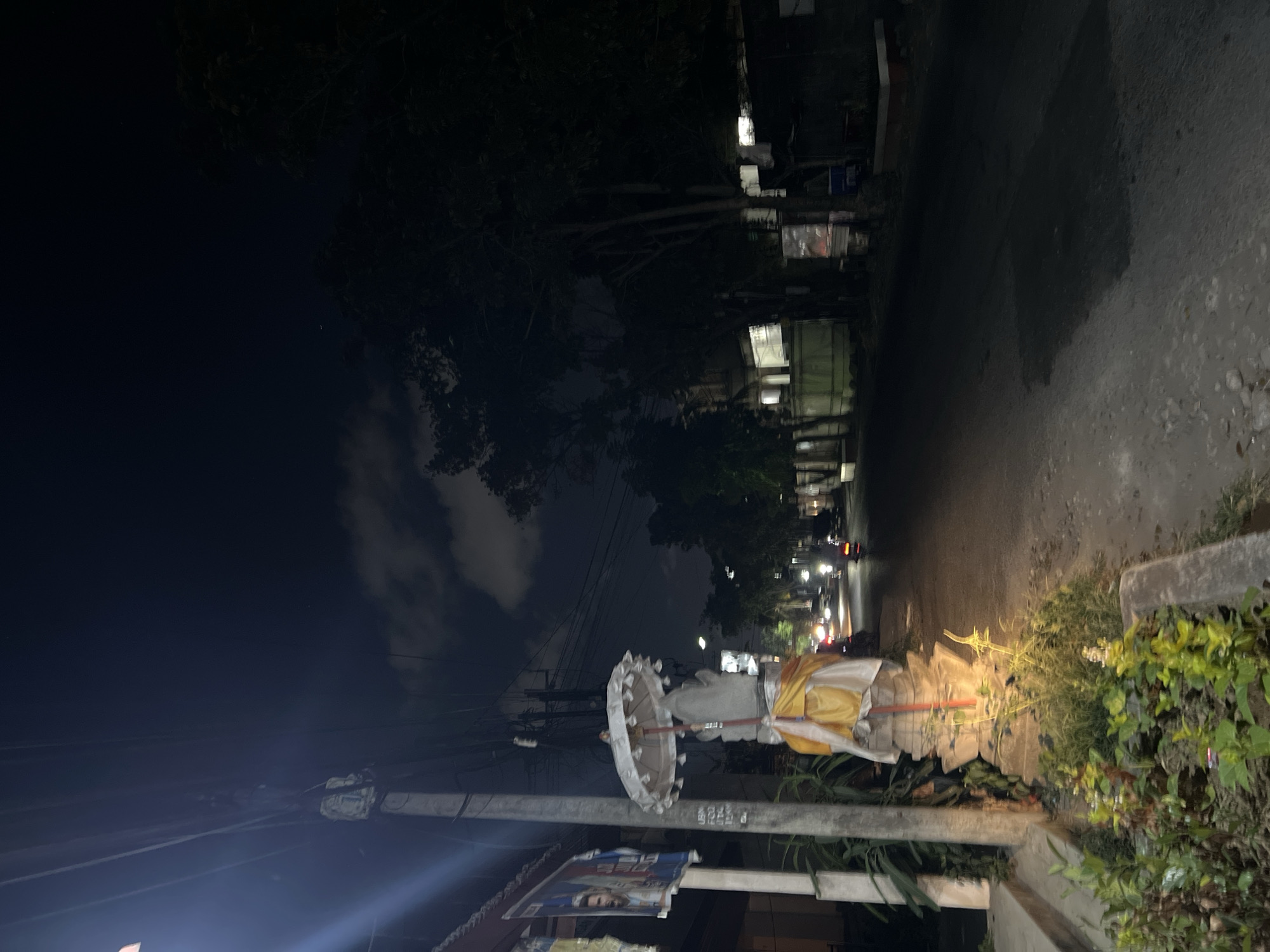 An eye-level, full outdoor shot captures a street scene at night in Denpasar, Indonesia, with artificial light sources illuminating the area. In the foreground, a stone statue, adorned with a yellow and white cloth and a decorative umbrella, stands on the left side of a paved road. The statue is surrounded by lush green vegetation. To the left of the statue, a utility pole with numerous electrical wires is visible, with posters attached to it. The road stretches into the distance, with sparse car and motorcycle traffic. Buildings line the right side of the road, some with lit windows, while dense trees with dark foliage create a shadowy backdrop against the night sky. A few scattered clouds are present in the dark expanse above.