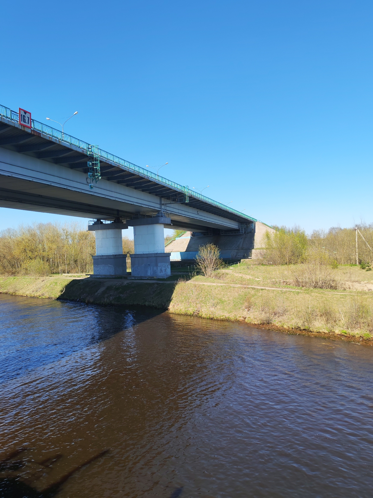 The image shows a bridge spanning a body of water, likely a river. The bridge is constructed with concrete supports and a gray, steel superstructure. A green railing lines the edge of the bridge, and a red and white sign with "16.7" is visible. The water is dark brown with ripples on the surface. The banks of the river are covered in grass and sparse vegetation. The sky is clear and blue.