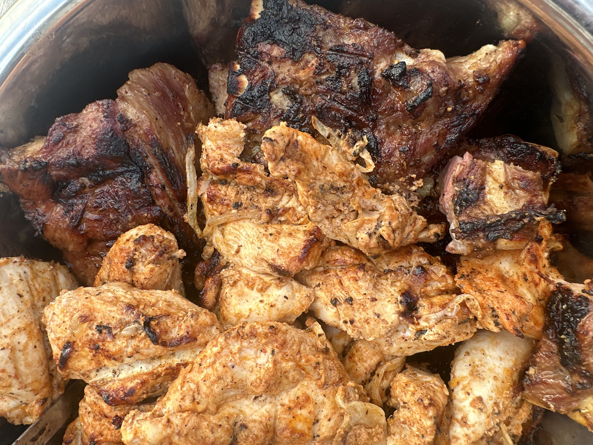 The image shows a close-up of a metal bowl filled with grilled meat. The meat consists of various cuts, including ribs and what appears to be chicken. Some pieces are heavily charred, while others are more lightly browned. The specific location in Chaikovsky, Russia, cannot be determined from this image. There are no people visible.