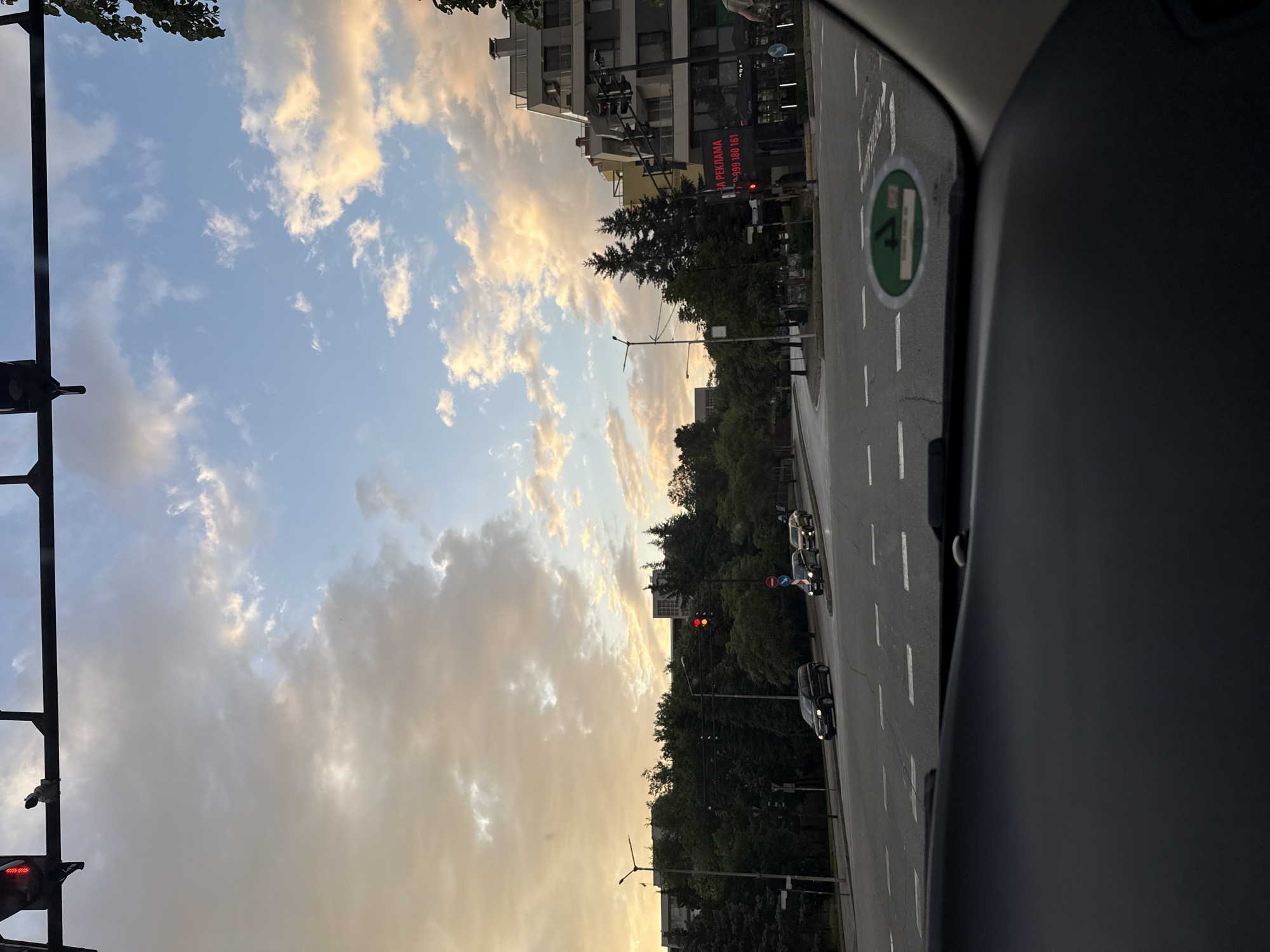 The image, taken from inside a vehicle, shows a street scene in Blagoevgrad, Bulgaria. The sky is filled with clouds, illuminated by the setting sun. A street runs vertically through the image, lined with trees. Buildings are visible on the left side of the street. Cars are stopped at a traffic light, which is currently red. A green environmental sticker is affixed to the windshield of the vehicle from which the photo was taken.