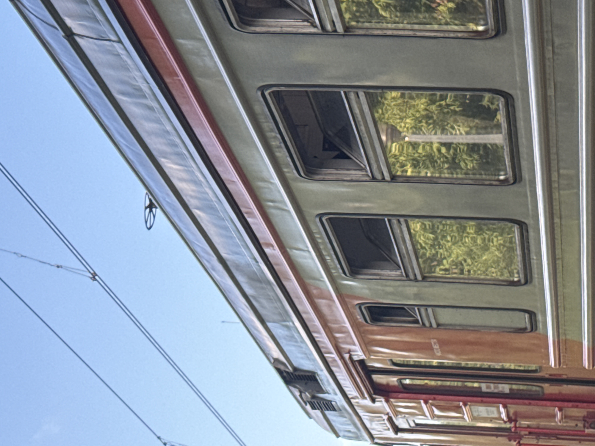 An exterior shot captures the side of a train car with a blue sky overhead. The train car is predominantly a muted green, with a reddish-orange section at the left end, featuring several doors and windows. The upper part of the train car has a darker, possibly metallic, gray roof with a red stripe running horizontally along its edge. Several windows are visible on the green section of the train, some reflecting a dense green foliage and a light pole. Overhead, a network of electrical wires, likely for the train, crisscrosses the sky. The image appears to be taken from a street-level perspective looking upwards at the train. The scene does not contain any people. The location is Yekaterinburg, Russia.