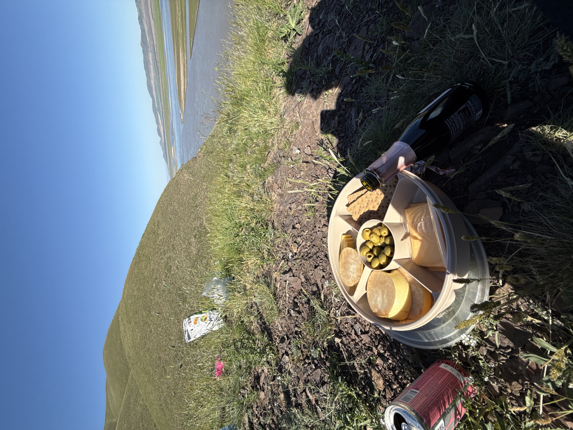 An outdoor scene captures a picnic setup on a sunny day in Ust'-Abakan, Russia. In the foreground, a circular, compartmentalized serving platter holds green olives, crackers, and several pieces of cheese, suggesting a light snack. A dark bottle, likely champagne or sparkling wine, with "PREMIUM" visible on its pink foil-wrapped neck, lies on the grassy and rocky ground next to the platter. Partially visible on the bottle's label are "750 ml" and "12%". A red aluminum can, identifiable by the word "ASAHI" on its side, is also on the ground. A person's shadow is cast on the rough terrain near the picnic items. Scattered small pieces of litter, including a white packet and a pink item, are visible on the ground. The background features a wide body of water, possibly a river or lake, reflecting the clear blue sky. Distant hills or low mountains, some with visible green fields, line the horizon. The bright sunlight and strong shadows indicate it is midday or early afternoon, with clear weather.