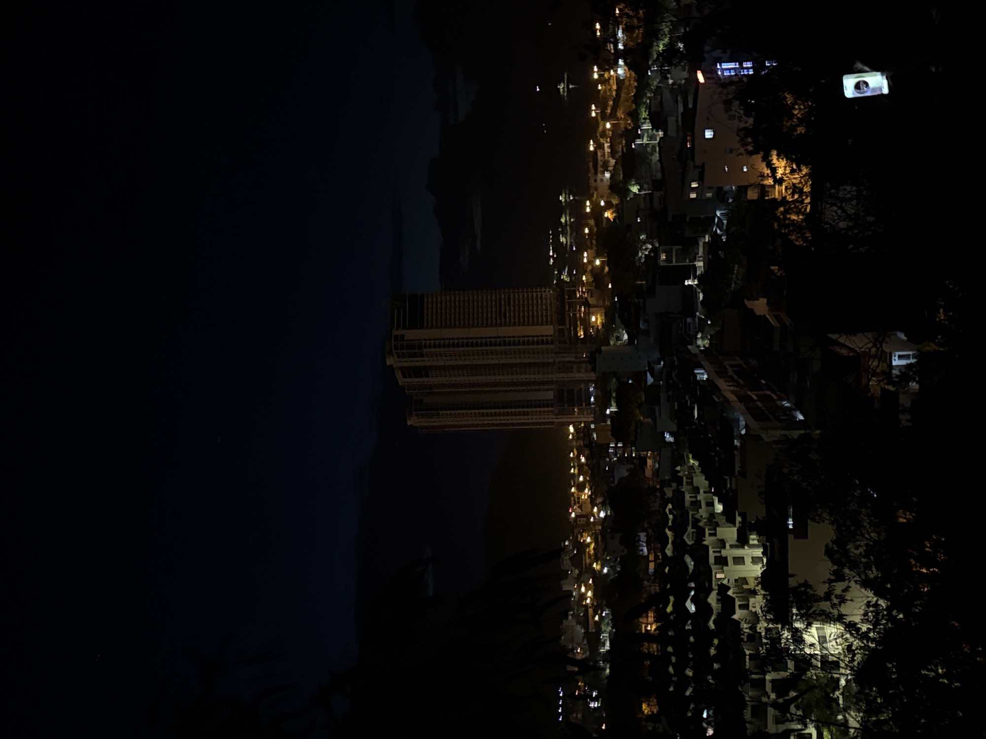 At night, a high-angle view shows the ward of Bac Nha Trang, Vietnam. A dark body of water stretches to the left, while the illuminated city extends to the right. Streetlights illuminate a road along the water. A large, modern building under construction stands out near the center. Urban development continues on the right side of the image. Trees line the right side.