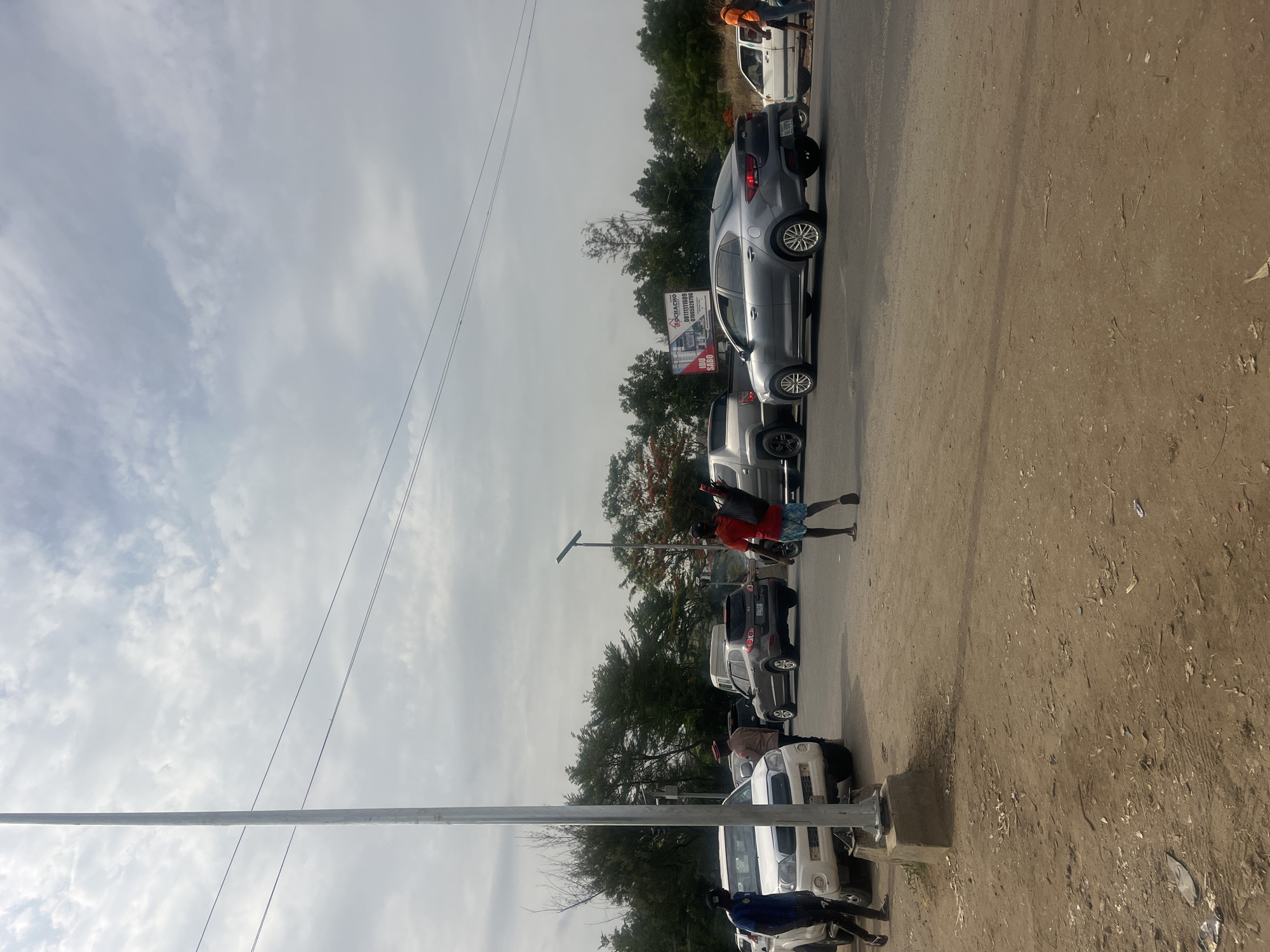 This media file captures an outdoor scene in Karum, Nigeria, depicting a roadside environment with parked vehicles and several people under an overcast sky.
A line of cars, including sedans and SUVs in shades of grey, silver, and white, are parked bumper-to-bumper along the side of what appears to be a paved road on the left, bordered by a wide, dusty, unpaved shoulder on the right. In the middle ground, a person wearing a red shirt and blue patterned shorts stands near a grey sedan, facing to the right. Further down the road, another individual in an orange vest and blue pants is visible near a white vehicle. Closer to the foreground on the left, a person in a blue top is partially visible next to a white SUV.
A prominent light-colored utility pole stands in the foreground on the left, with thin overhead wires stretching across the frame against the cloudy sky. In the background, behind the parked vehicles and some scattered trees, a billboard is partially visible. Text on the billboard includes "D-GHACRO," "CONSTRUCTION," a phone number starting with "070," and "IBD SABO." The ground on the right side of the image is barren, dusty earth with some scattered debris. The sky is largely cloudy or overcast, indicating daytime with diffuse lighting and no strong shadows. The scene suggests a typical roadside or street activity, possibly traffic or vehicles parked along a route.