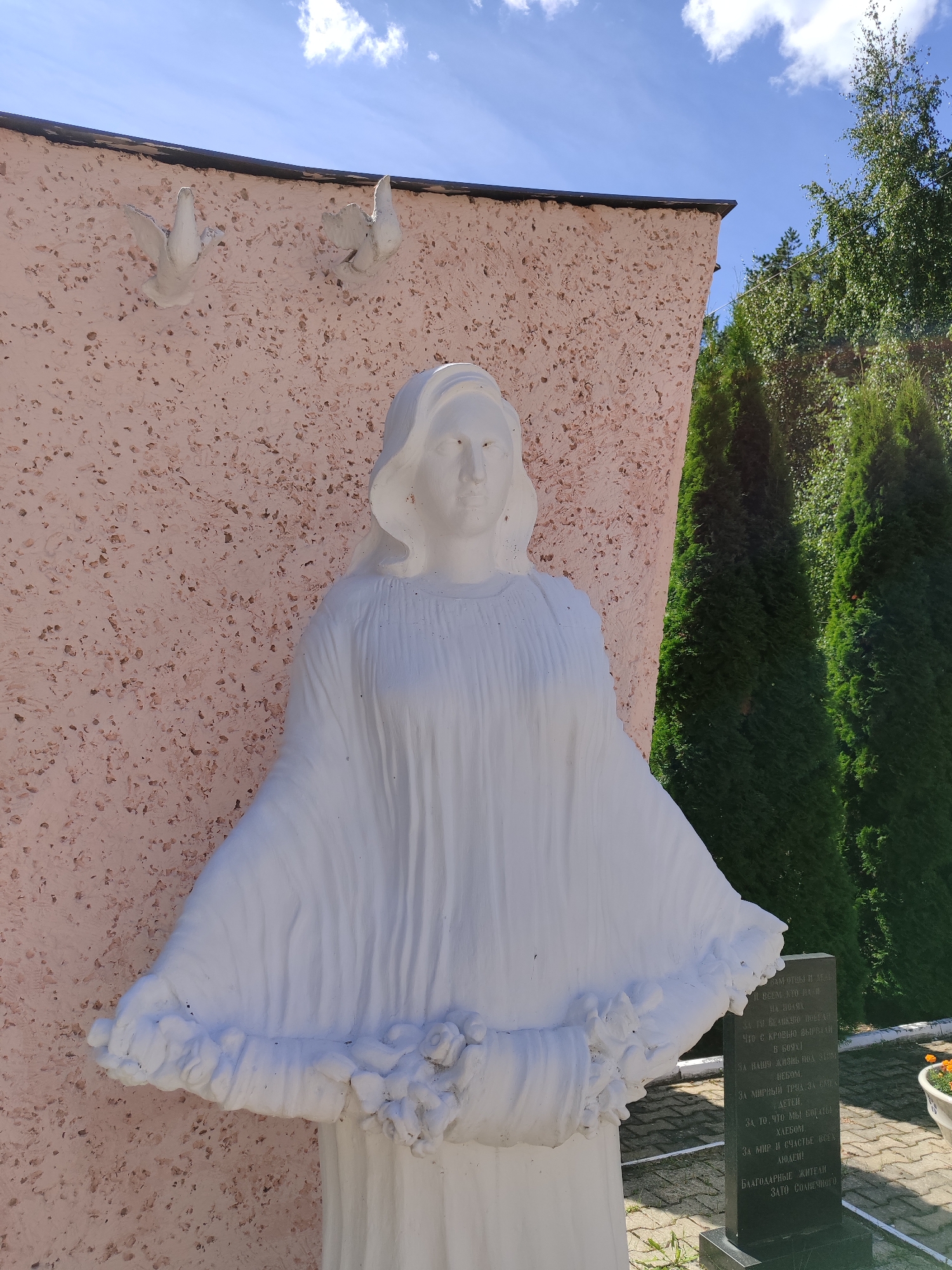A white statue of a woman stands in front of a pink, textured wall. The statue is draped in flowing robes and holds a wreath of flowers at her waist. Two white bird sculptures are attached to the wall above and to the left of the statue. To the right of the statue, a black tombstone with white text is visible. Green coniferous trees and a bright blue sky with white clouds form the background. The ground is paved with gray tiles, and a white planter with orange flowers is partially visible in the lower right corner.