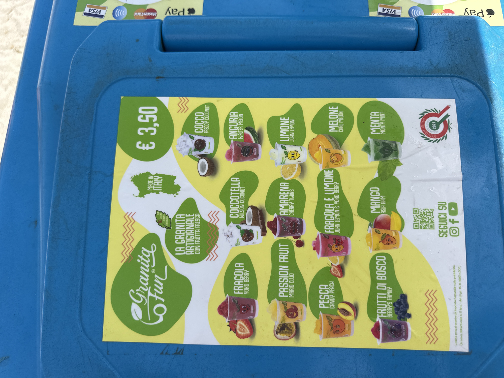 The image shows a bright yellow menu board advertising "Granita Fun" drinks, affixed to a textured blue plastic surface, likely a cooler or kiosk. The menu features various fruit flavors like strawberry, coconut, passion fruit, cherry, lemon, peach, strawberry and lemon, berry's family, mango, mint, and melon, each with a playful cartoon character in its cup. The price is listed as € 3.50. The text indicates the granita is artisanal and made with fresh fruit, and "Made in Italy." Payment icons for Visa and Mastercard are visible on the right side of the blue surface, along with the words "Pay" and an Apple Pay logo. A QR code and social media icons (Instagram, Facebook, YouTube) encourage customers to "Seguici Su" (Follow us). A seal at the bottom left indicates "Eccellenza Italiana 2021." The background is out of focus but appears to be outdoors, possibly a beach or tourist area, given the context of "Crabonaxa/Villasimius, Italy" which is a coastal area in Sardinia known for tourism. The image does not contain any people.