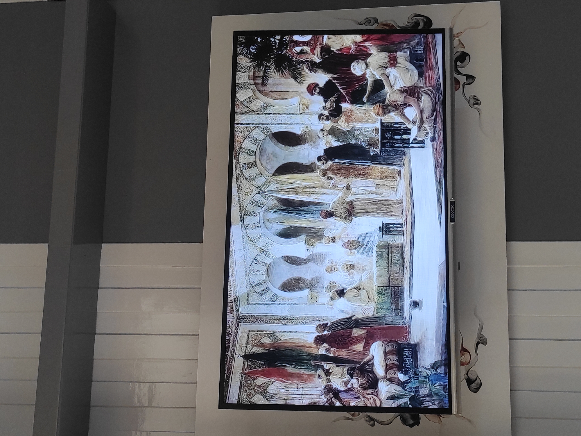 This image shows a flat-screen television displaying a painting. The painting depicts a gathering of men in a room with arched doorways and ornate walls, suggesting an interior architectural setting. The men are dressed in traditional attire, including turbans and robes, and appear to be engaged in conversation or discussion. Some men are seated on cushions, while others are standing. The overall scene is rich in detail and color, conveying a sense of historical or cultural significance. The television is mounted on a wall which has a textured, segmented pattern. The image itself does not provide any specific information about the city of Sumedang, Indonesia.