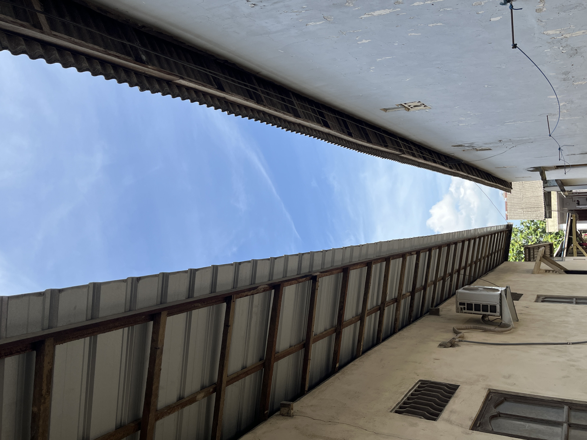 The image captures a view looking upwards between two buildings, with a bright blue sky and scattered clouds overhead. On the left, a light-colored building with a light gray corrugated metal roof is visible. The roof is supported by wooden beams. An air conditioning unit is mounted on the wall of this building, below the roofline. A window with a metal grate is also visible on this side.
On the right, a light blue building with a weathered, textured facade is seen. This building has a darker, corrugated roof made of what appears to be asbestos or similar material, also supported by wooden beams. Two small, decorative windows are set into the wall of this building. A dark wire runs horizontally across the wall, secured by blue string.
In the background, hints of other buildings and lush green trees can be seen, suggesting a residential or semi-urban environment. The scene is likely from the city of Sejahtera, Indonesia, given the architectural style and tropical foliage. There are no people or animals present in the image, and no discernable actions or interactions are taking place. The overall impression is one of a simple, perhaps older, urban dwelling area on a clear day.