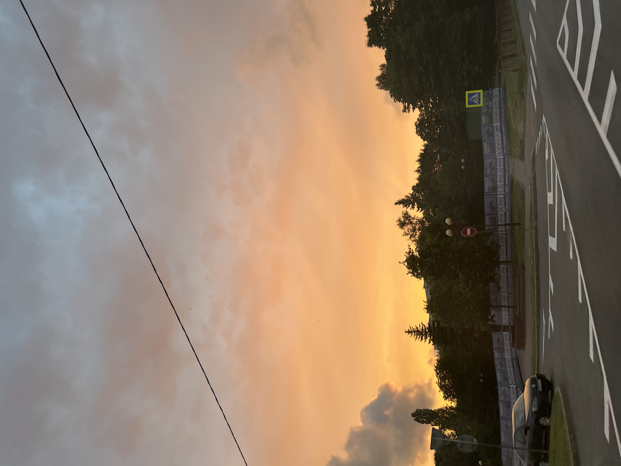 The image depicts a street scene in Minsk, Belarus, during what appears to be sunset or a heavily overcast day, as evidenced by the warm, orange glow in the sky and the soft lighting. The sky is filled with dramatic clouds, transitioning from a pale grey on the left to vibrant hues of orange and yellow towards the right, suggesting the sun is low on the horizon or obscured by clouds.
In the foreground, a dark-colored car is parked on the side of a road. The road itself is marked with white pedestrian crossing lines, indicating a crosswalk. To the left of the road, a sidewalk or pedestrian area is visible, bordered by a decorative fence with intricate blue and white patterns. Behind the fence, numerous trees create a dense, green backdrop, with some taller trees and evergreens interspersed. A yellow road sign with a blue symbol (likely a pedestrian crossing warning) is also visible on the left side of the scene, near the fence.
A thin black wire, possibly a utility line, stretches diagonally across the upper left portion of the frame, from the bottom left corner upwards. There are no visible people in the image. The overall mood is tranquil and perhaps a little melancholic due to the diffused light and the quiet street. The presence of the pedestrian crossing and the decorative fence are subtle cues to the urban environment of Minsk. The time of day is definitively late afternoon or evening due to the sky's coloration.