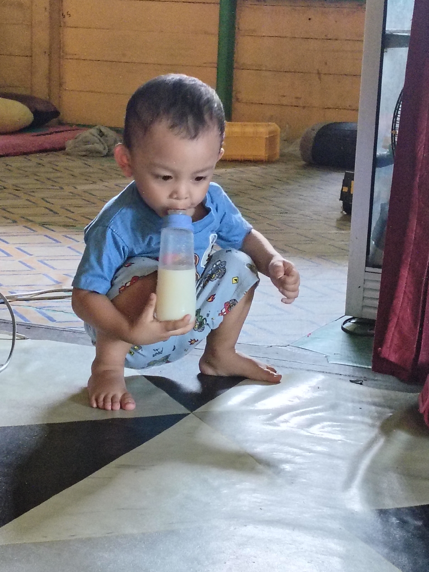 A young child is crouched on a tiled floor, drinking from a clear plastic bottle filled with a pale liquid. The child is wearing a blue t-shirt and patterned pants. The floor is tiled with a geometric pattern of black and white squares. In the background, there is a wooden wall and some furniture, including a yellow plastic box and a dark circular object. To the right of the child, a red curtain hangs beside a white structure that appears to be a door or window frame. The overall scene suggests an indoor domestic setting in Tanah Grogot, Indonesia.
