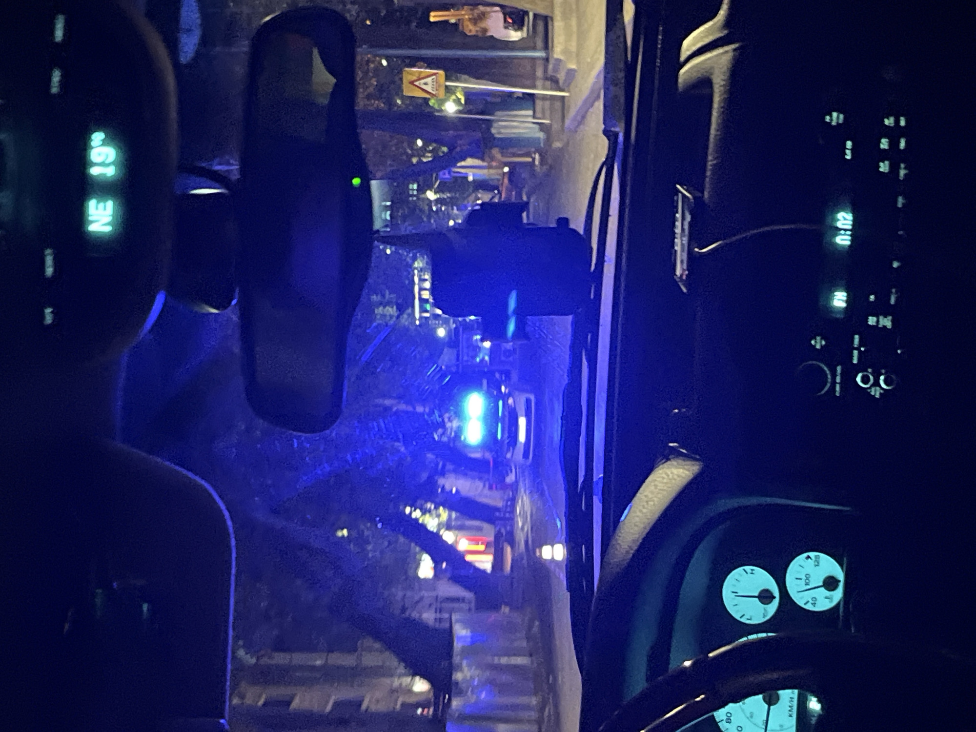 The image is taken from inside a vehicle, likely a car, at night. The digital display shows "NE 19°", and a clock displays "01:02". Outside the windshield, a street scene is visible, with trees and building lights. There's a vehicle with blue lights in the distance. A traffic sign can be seen with the text facing upside-down. Without other information, it's not possible to definitively place this location within Plovdiv, Bulgaria.