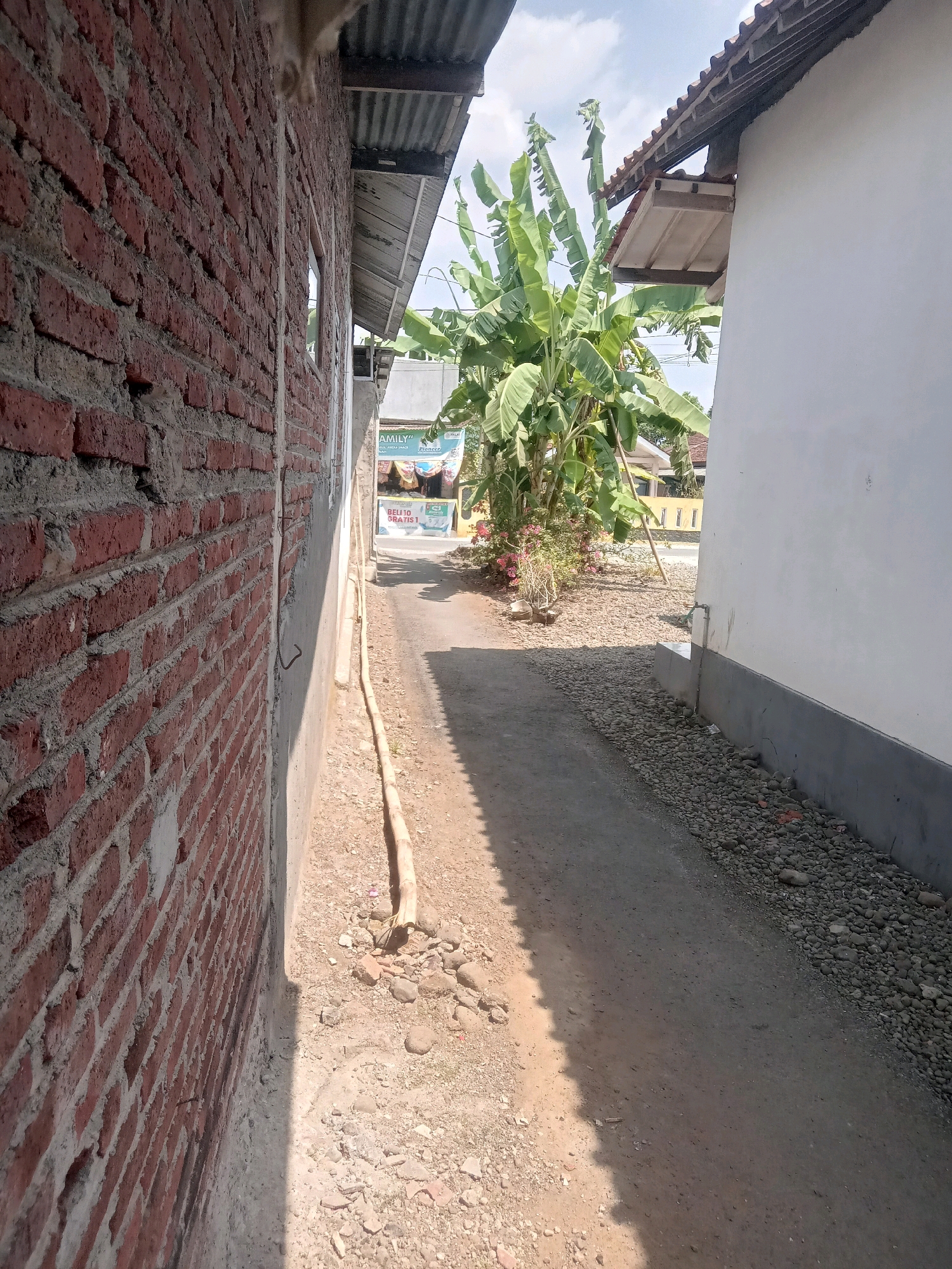 The image depicts a narrow alleyway between two buildings in Desa Klahang, Indonesia. On the left, a rough brick wall is visible, partially covered by concrete. To the right, a white wall with a dark grey base runs along the alley. The ground is a mixture of dirt and gravel, with a yellow pipe running along the wall. In the background, a banana tree with large leaves stands tall, and further back, a building with a tiled roof and yellow window frames can be seen. Sunlight casts a shadow across the alley. There are no people present in the image.